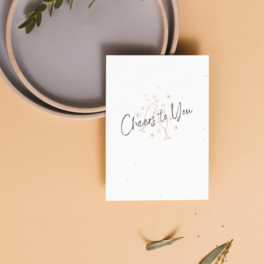 White card with 'Cheers to You' text on a beige surface with a gray plate and green leaves.