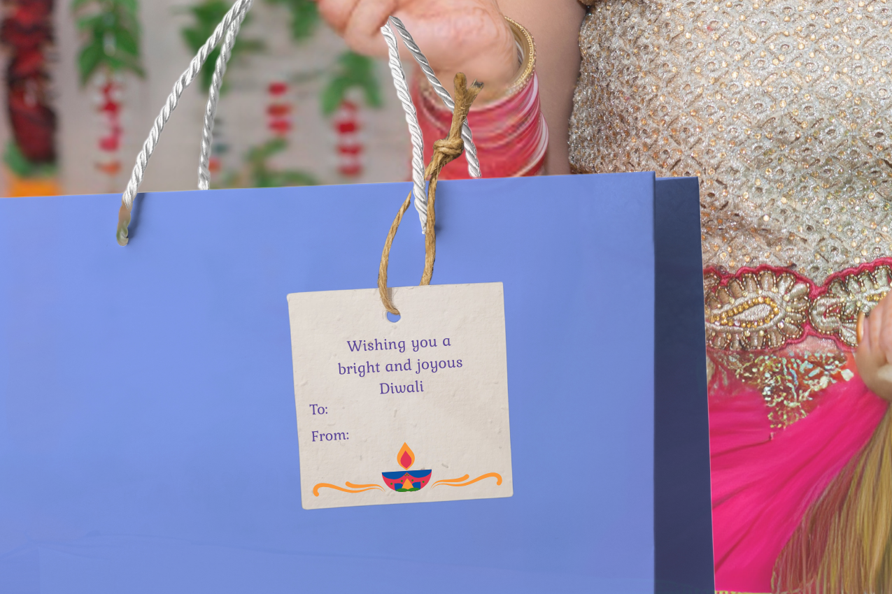 Woman holding a blue gift bag with a 'diya' tag with a message for a 'broght and joyous Diwali', against a colorful background