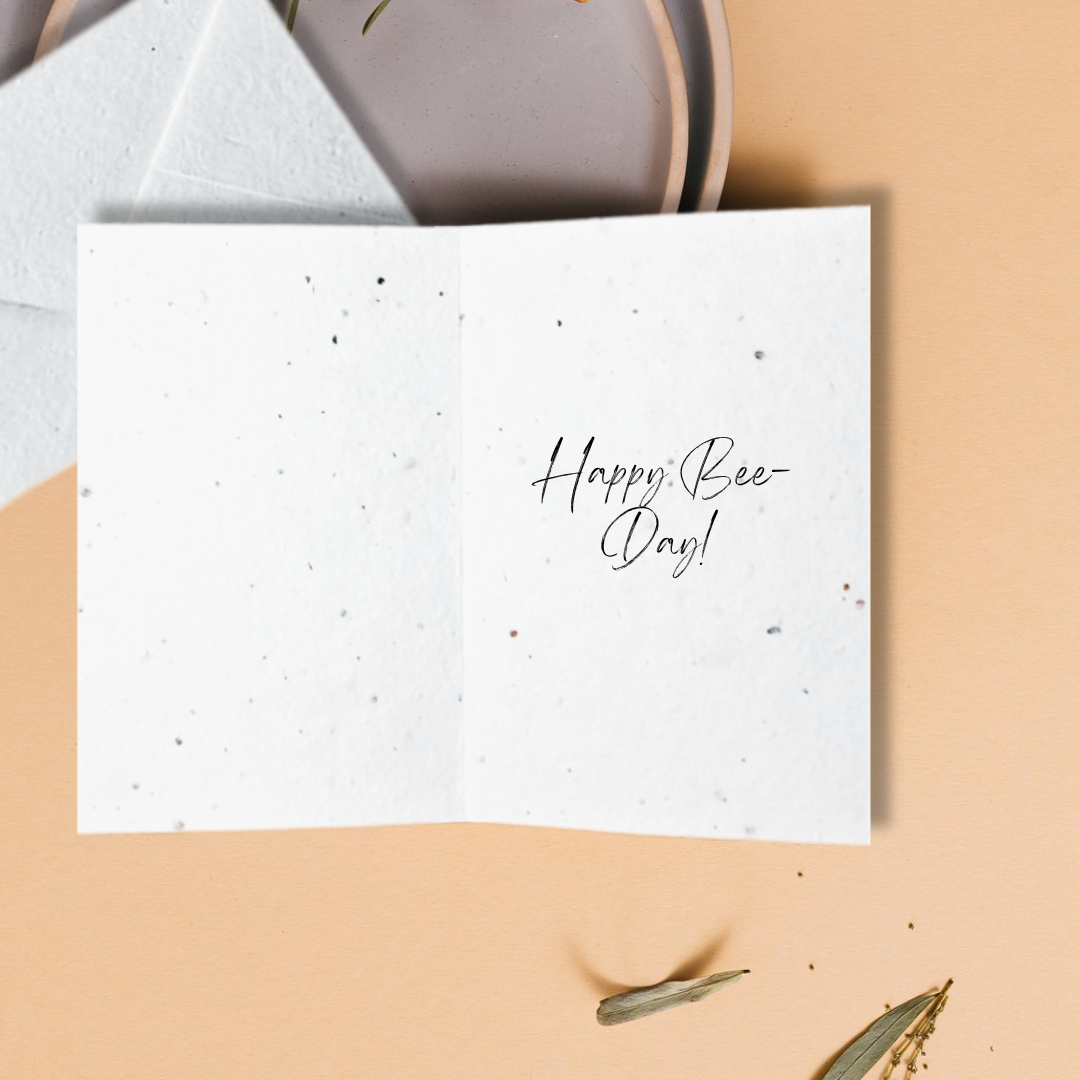 White card with 'Happy Bee Day!' text on a beige surface with envelopes.