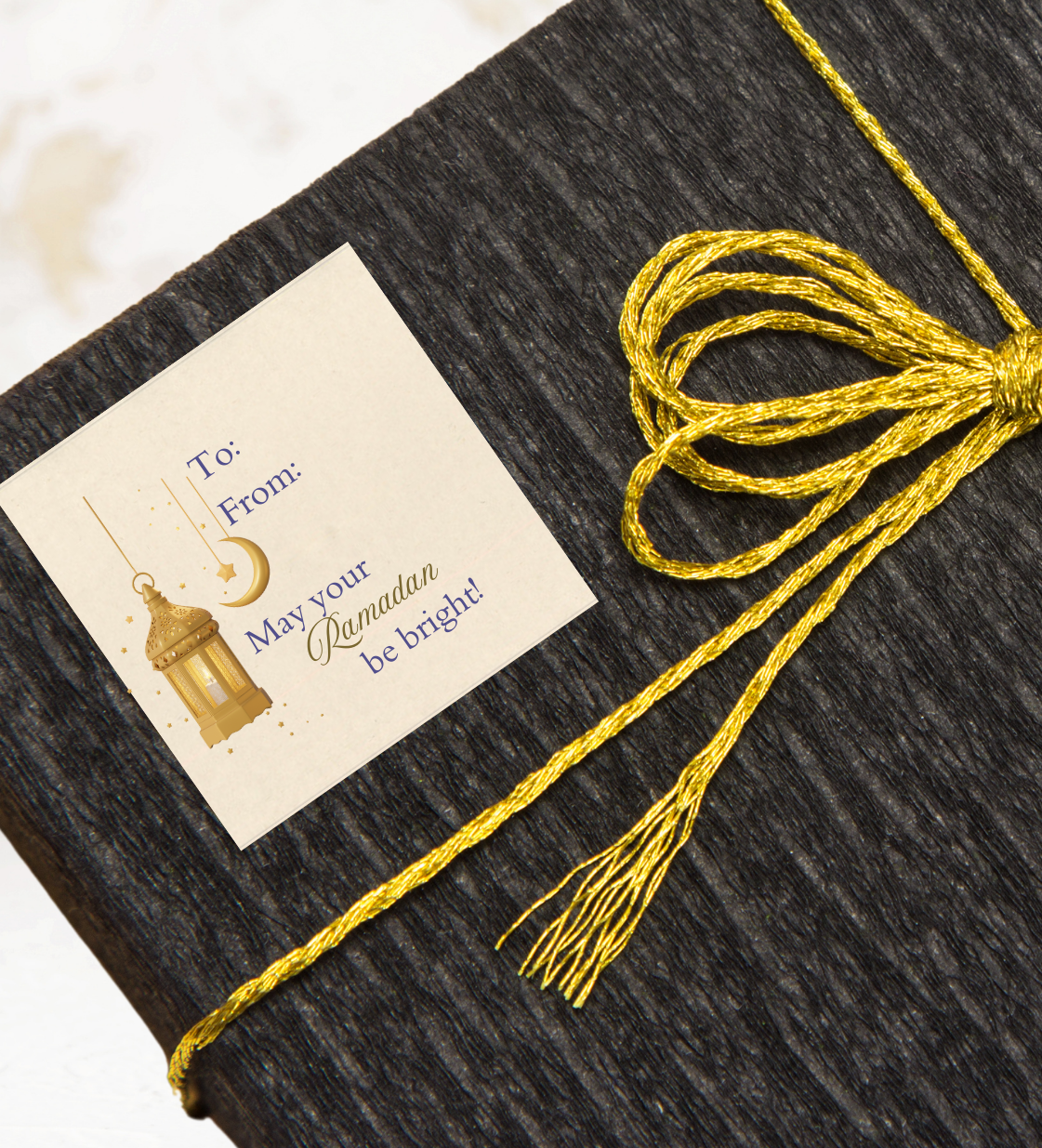 Dark textured gift box with a gold ribbon and a decorative gift sticker label with 'May your Ramadan be Bright' text on a light background