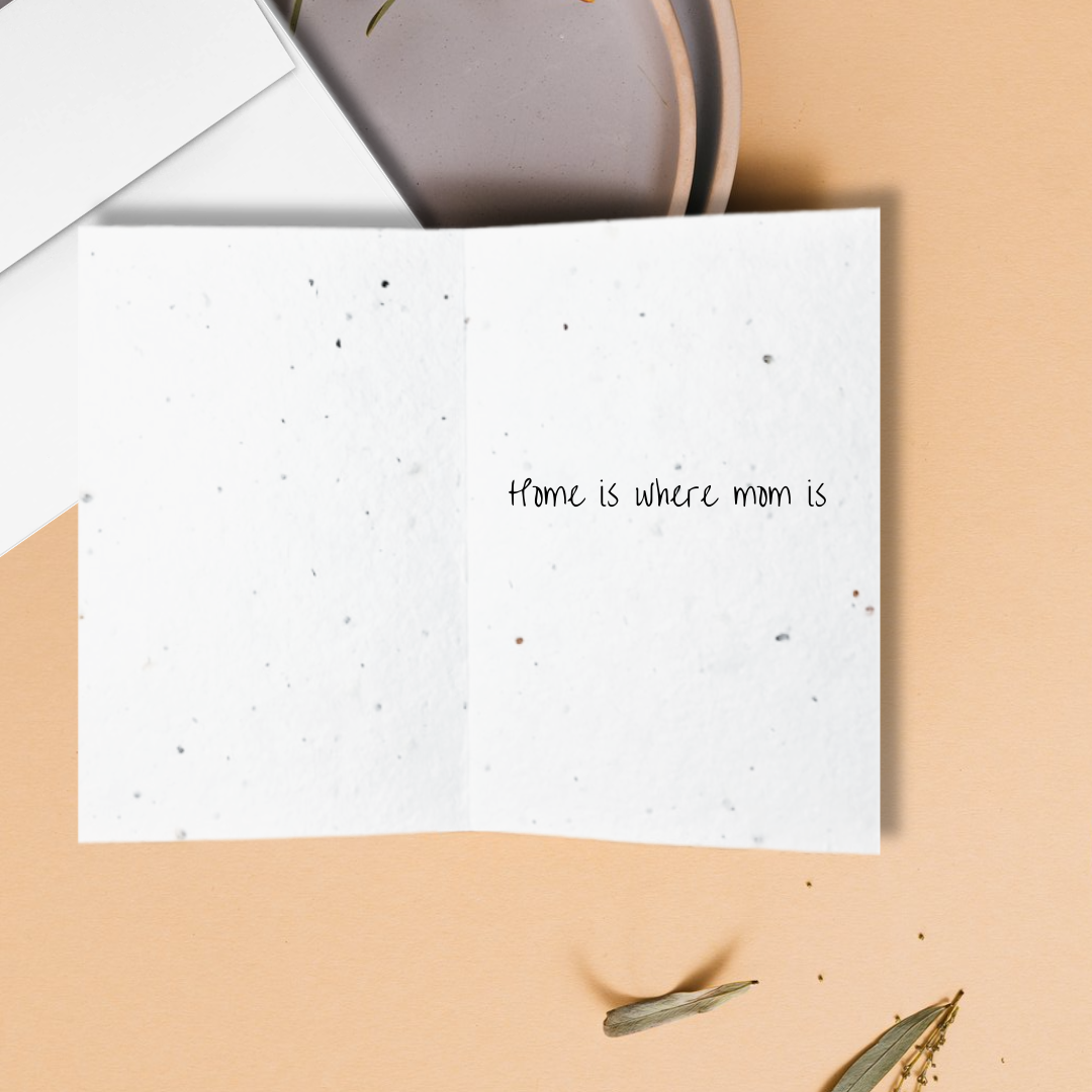 Mother's day card with 'home is where mom is' text on a beige surface with a white envelope and a brown ceramic item.  For Mother's day