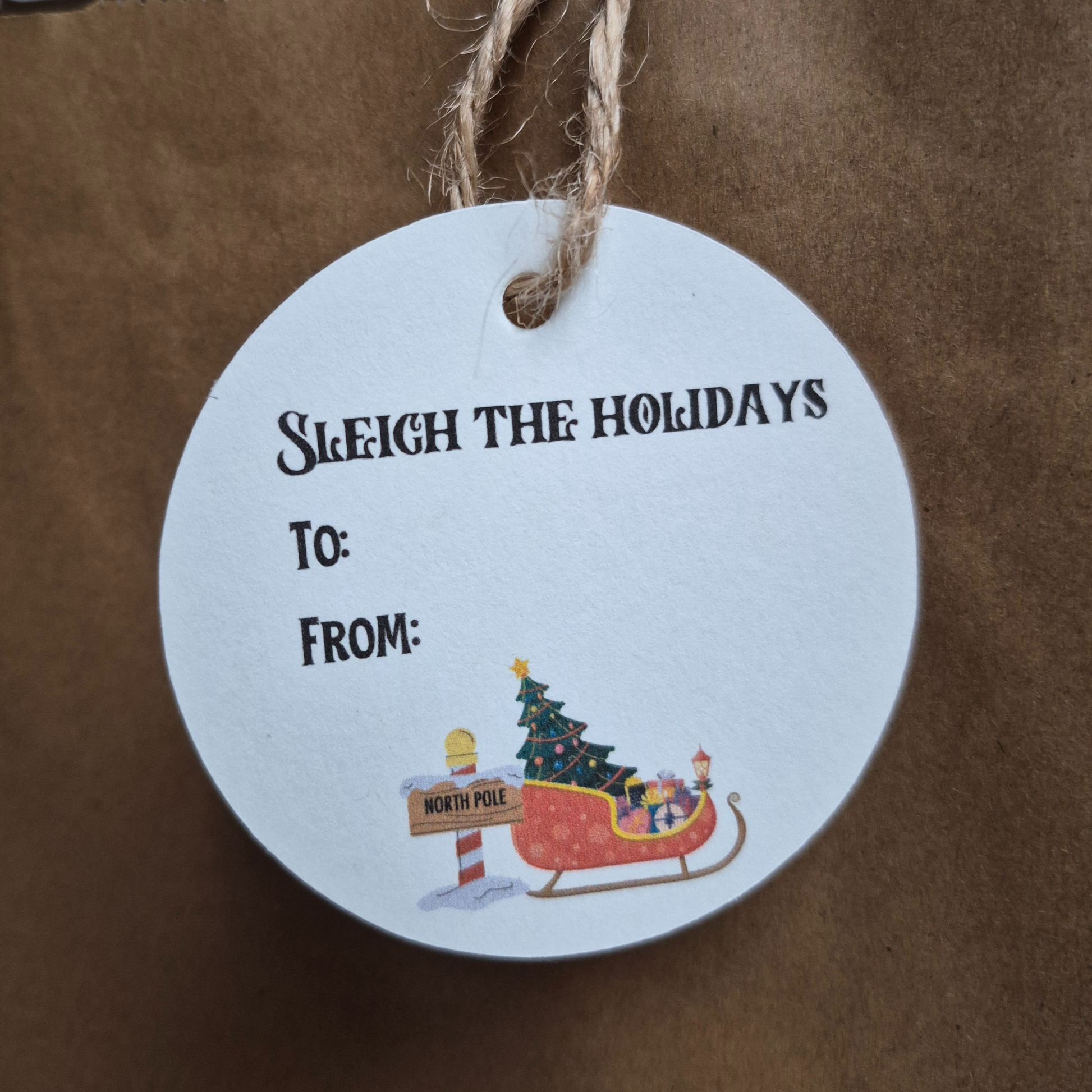 Round Recycled paper Christmas gift tag with 'Sleigh the Holidays' text and Santa's sleigh illustration on a brown paper bag background.