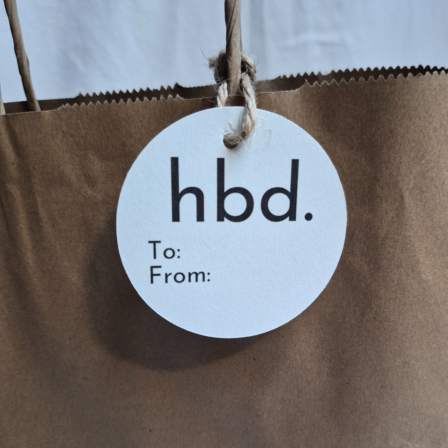 Round Birthday gift tag made of recycled paper with minimalist 'hbd.' text on a brown paper bag.