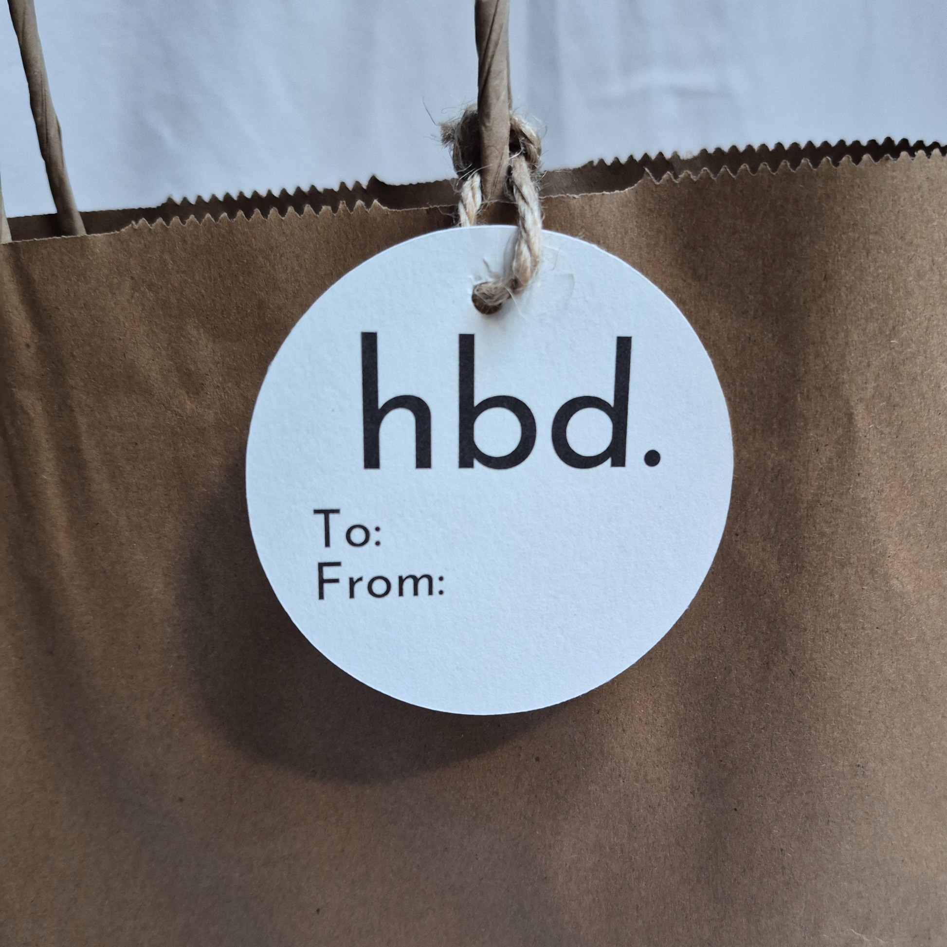 Round Birthday gift tag made of recycled paper with minimalist 'hbd.' text on a brown paper bag.