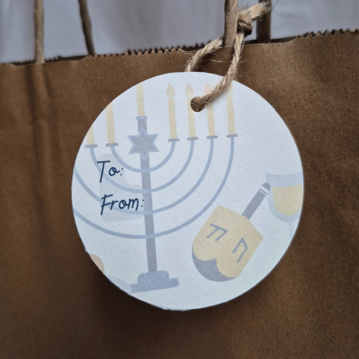 Round Hanukkah Gift tag made of recycled paper with Menorah and dreidel design. Hanging from a brown paper bag