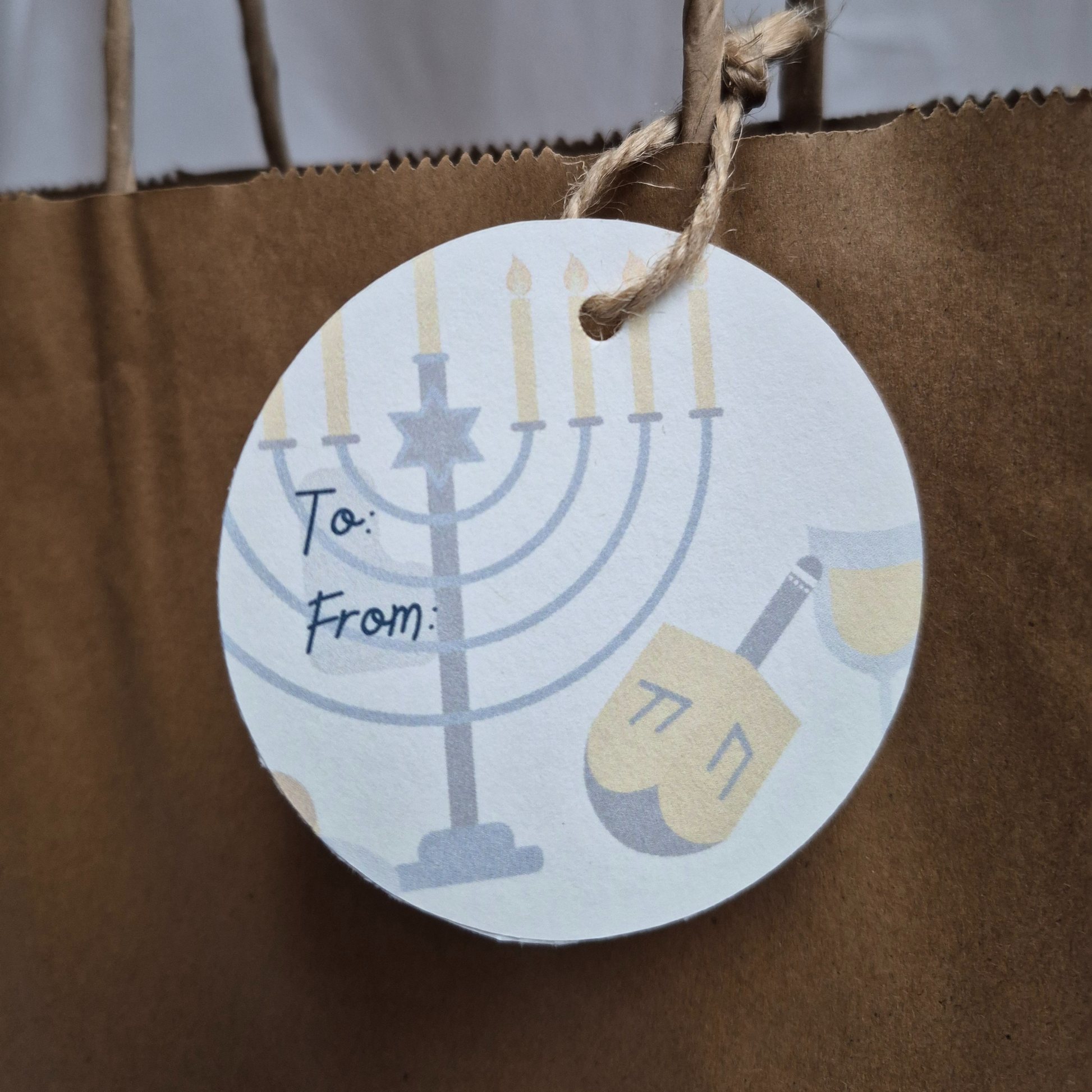 Round Hanukkah Gift tag made of recycled paper with Menorah and dreidel design. Hanging from a brown paper bag