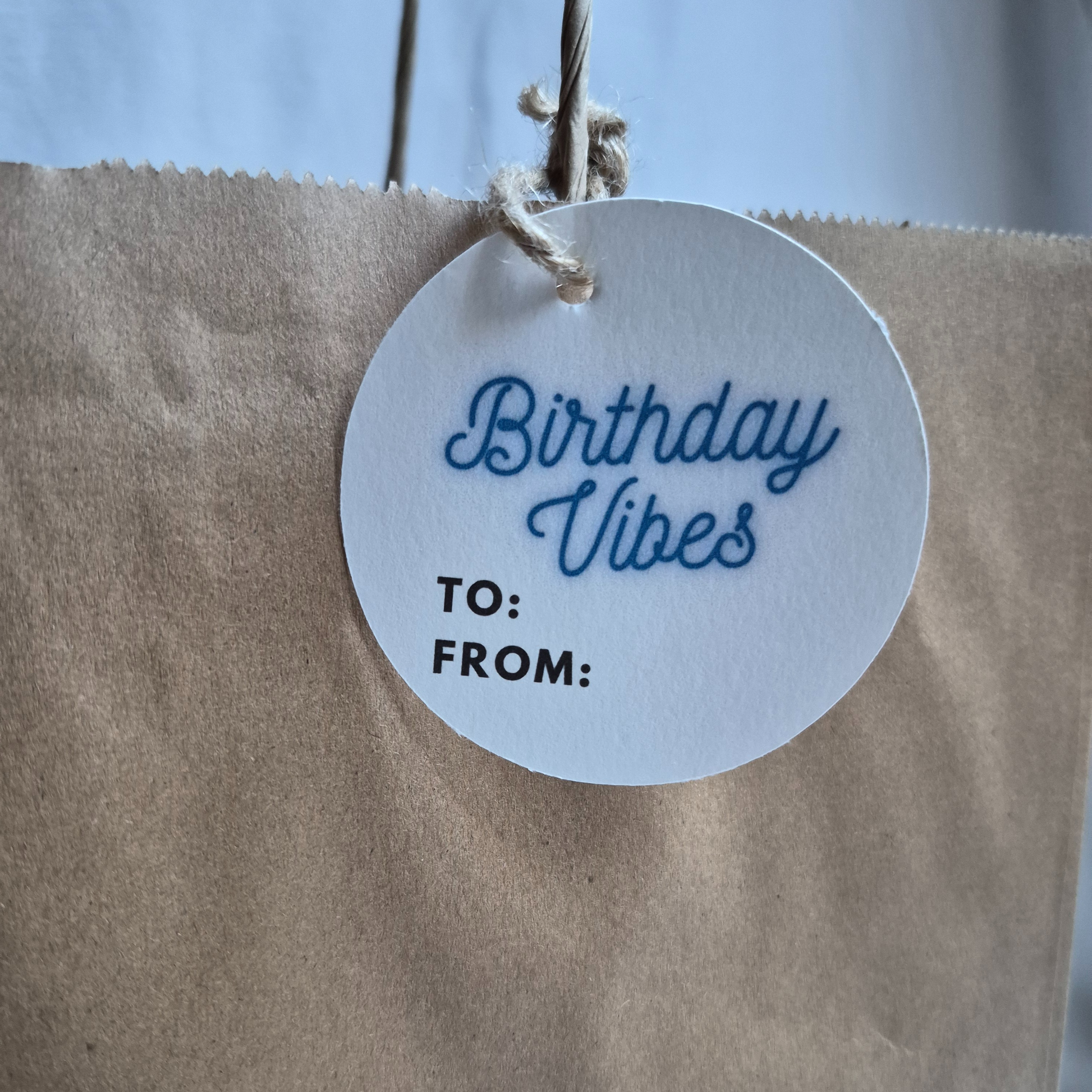 Round Birthday gift tag made of recycled paper with 'Birthday Vibes' text in neon blue on a brown paper bag