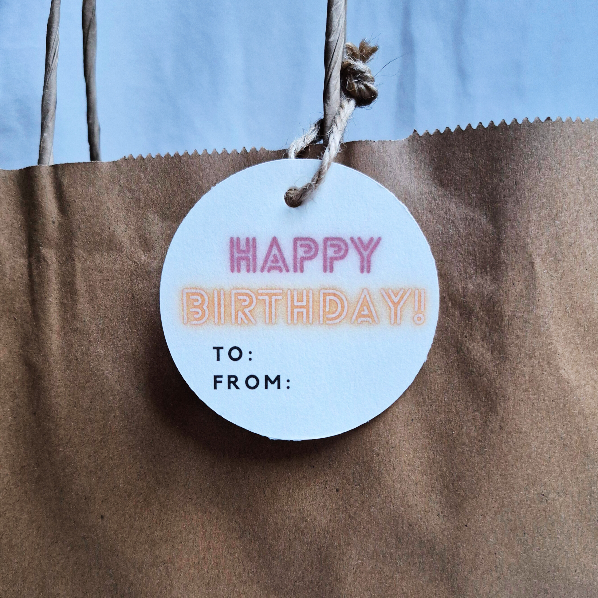 Round birthday tag made of recycled paper with 'Happy BIrthday' in neon text on a brown paper bag