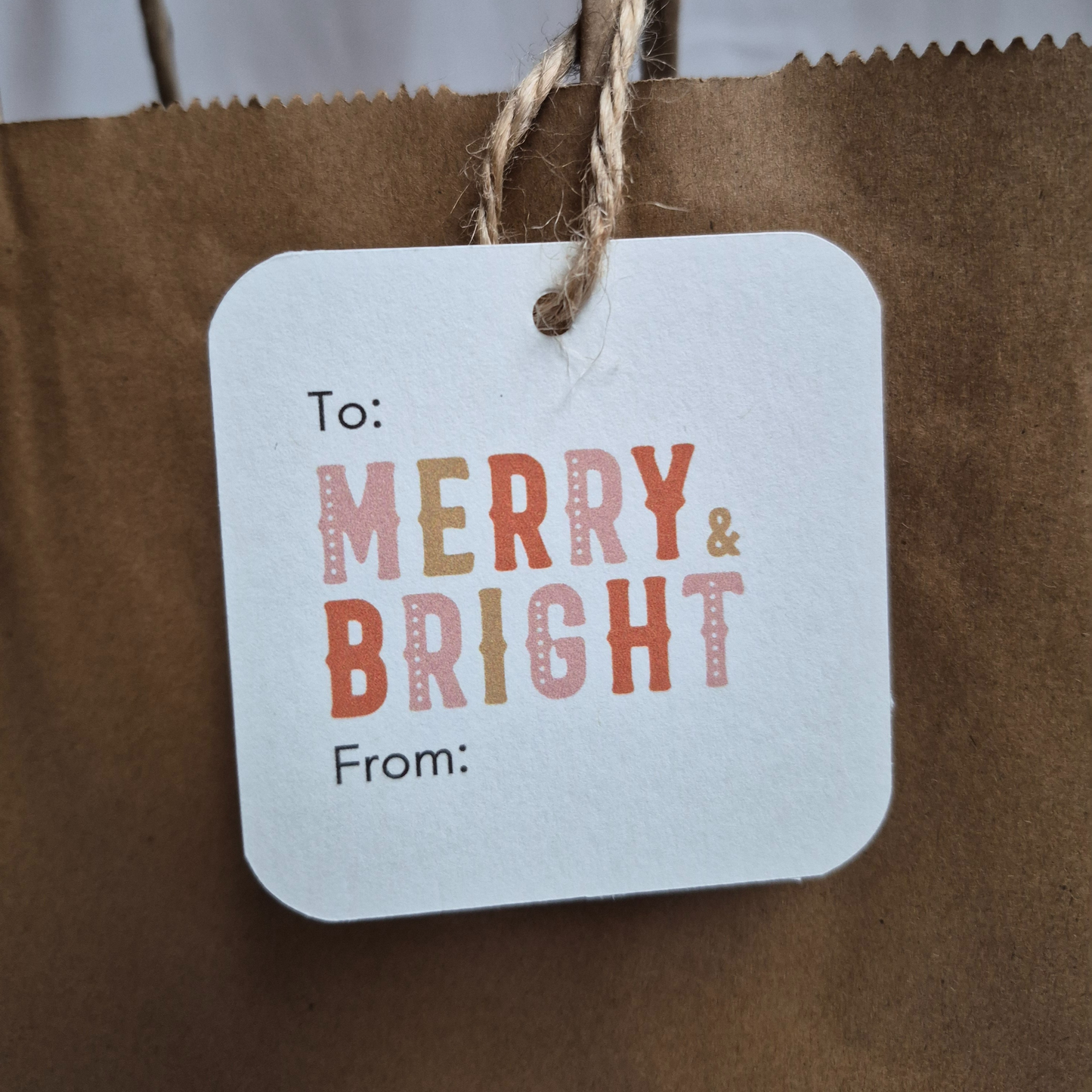 Square recycled paper Christmas gift tag with 'Merry & Bright' text hanging on a brown paper bag.