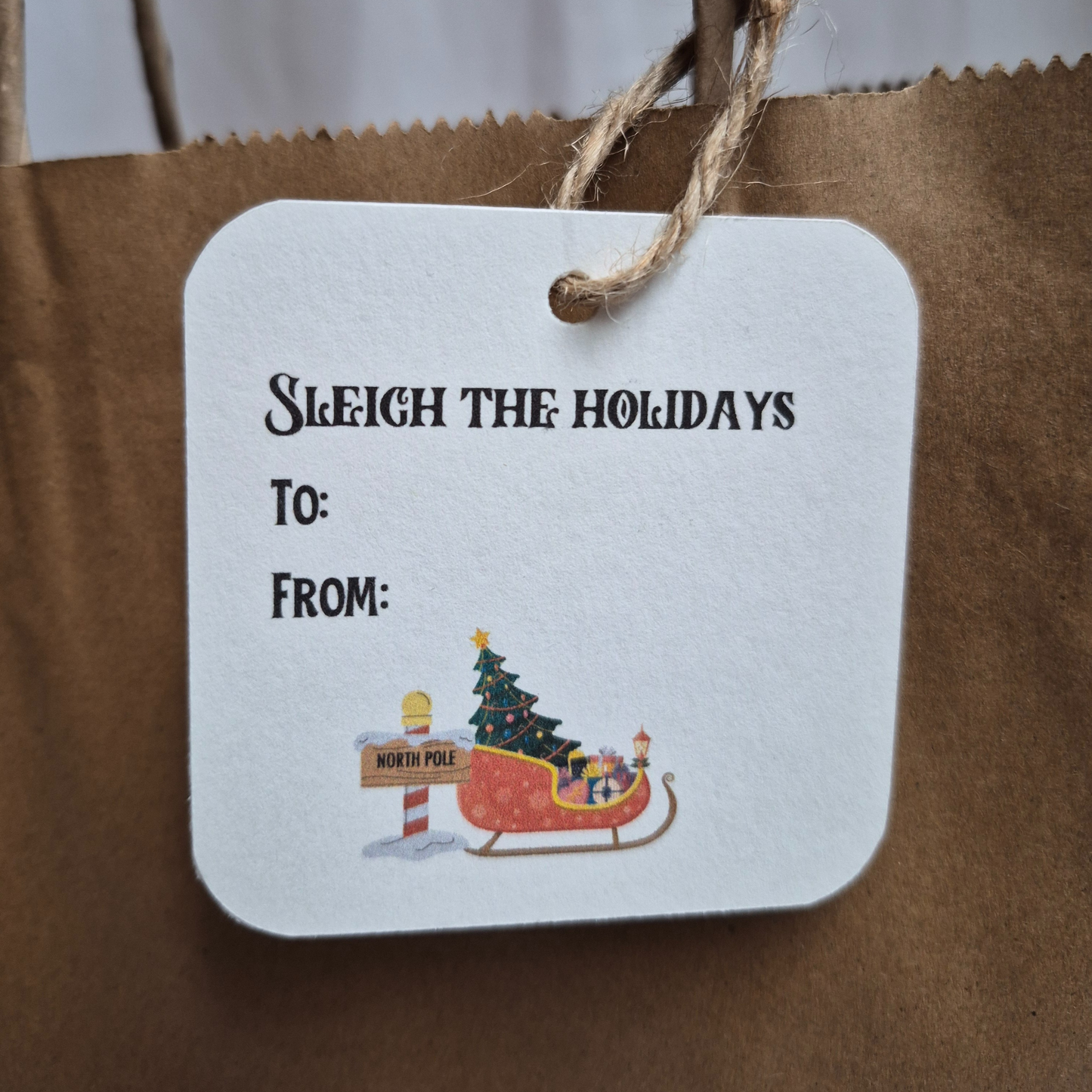 Square Christmas gift tag made of recycled paper with 'sleigh the holidays' text and sleigh and tree deign on a brown paper bag.