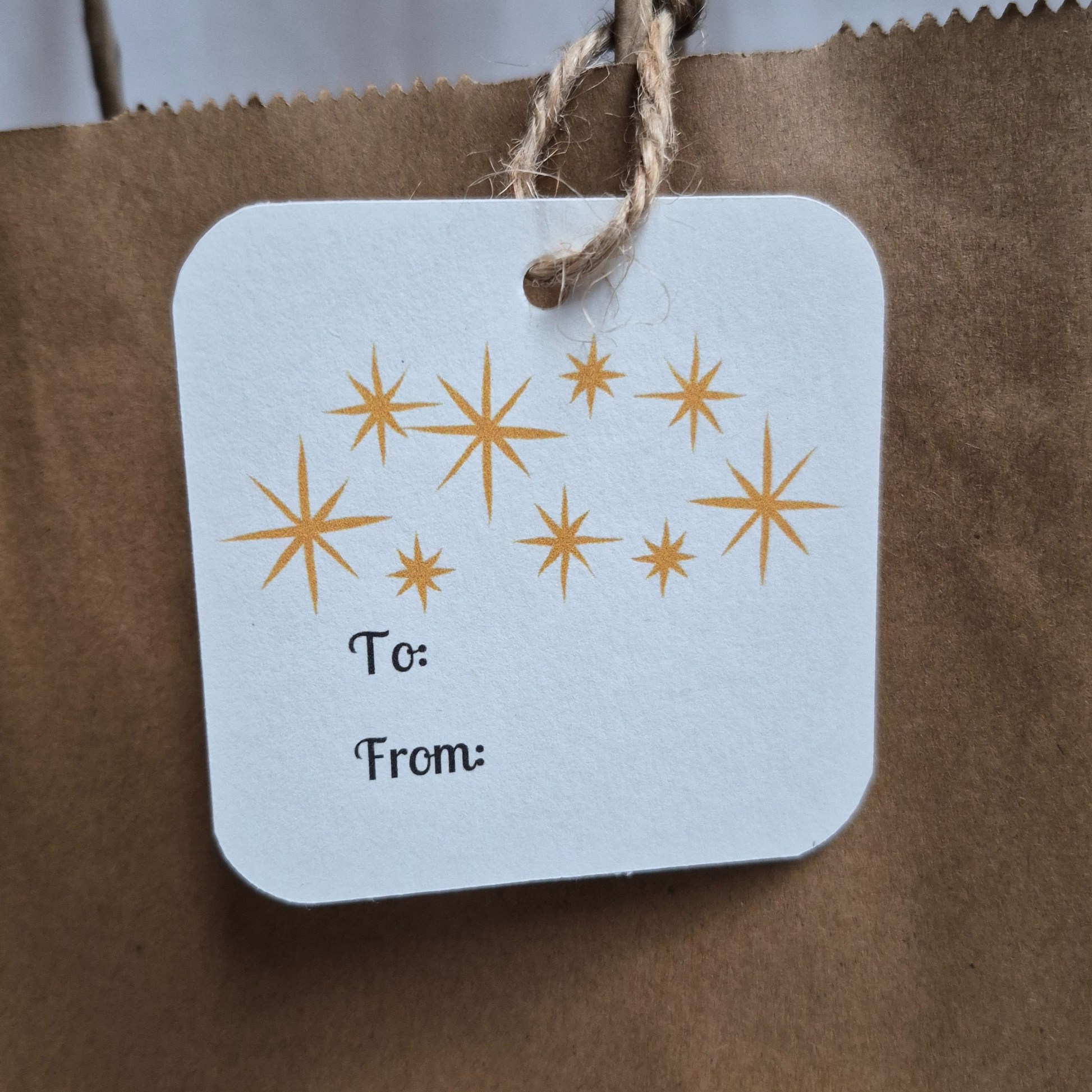 Square recycled paper Christmas Gift tag with gold stars and 'To:' and 'From:' text on a brown paper bag.