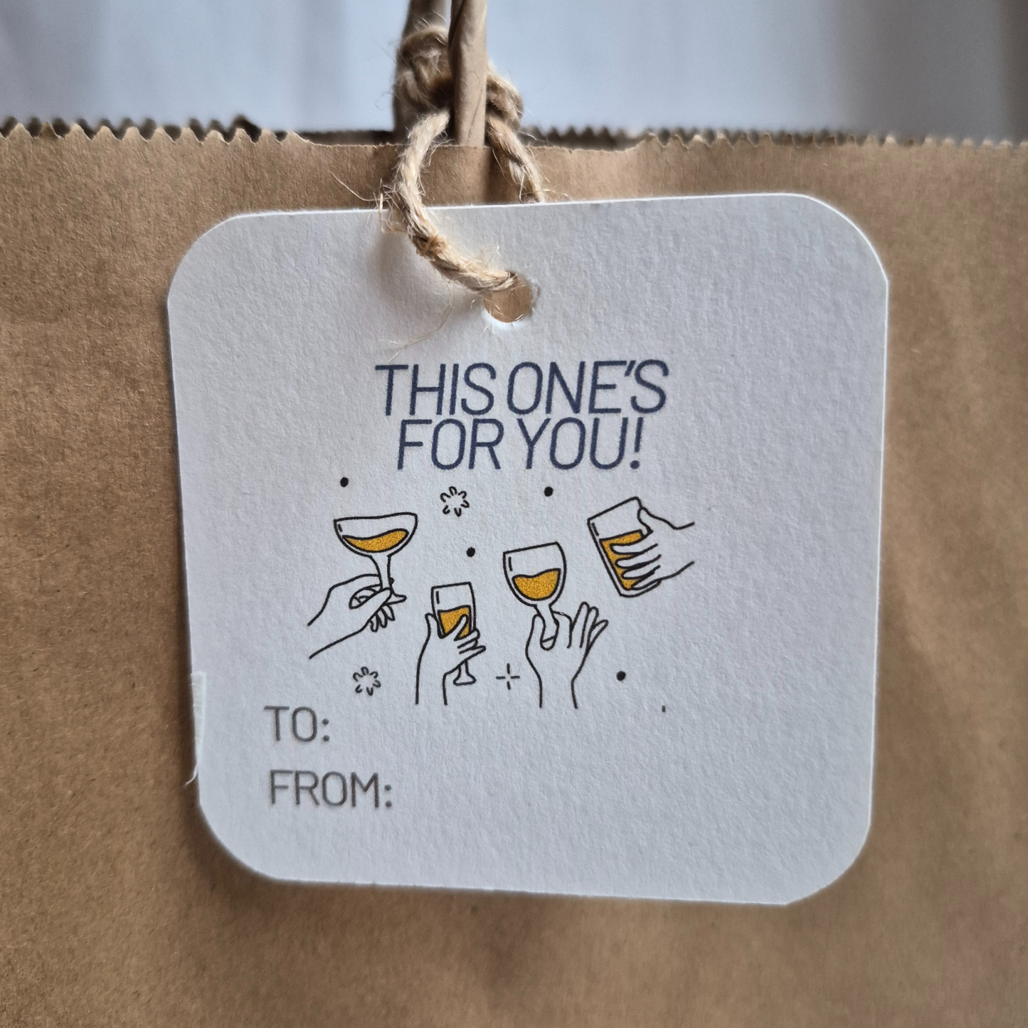 Square recycled paper gift tag with 'This one's for you!' text and wine glass illustrations on a brown paper bag.