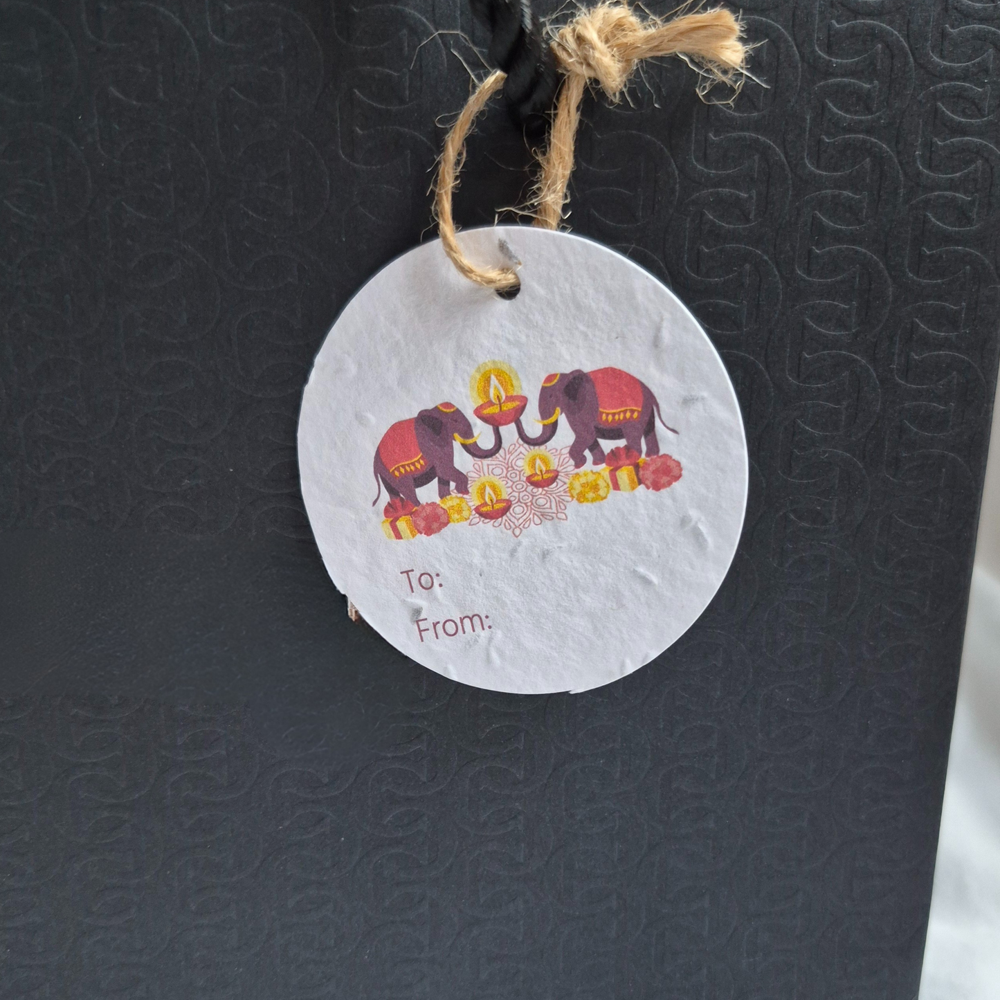 Round Diwali gift tag, made of seed paper, with elephants and diyas illustration hanging from a black bag