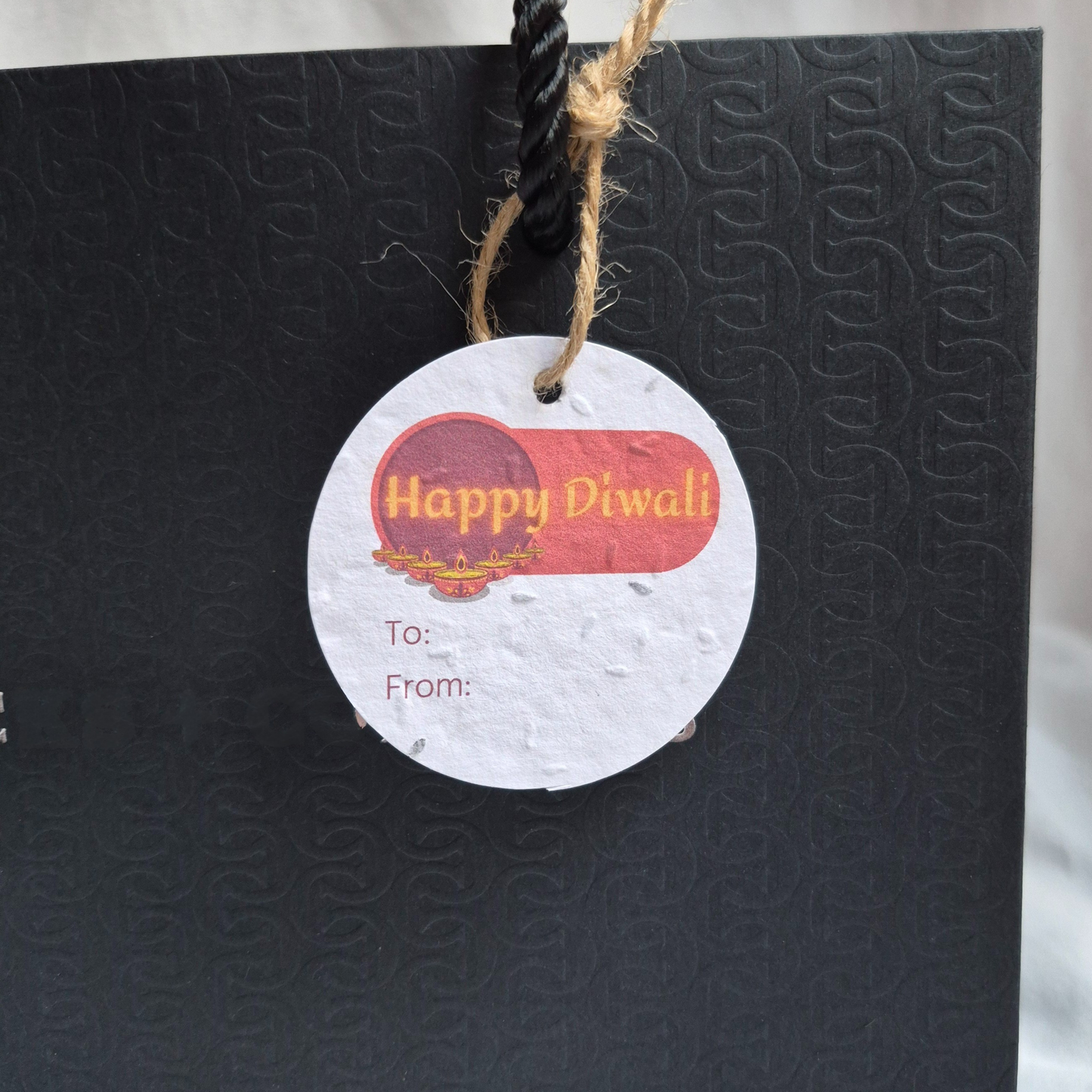 Round Diwali gift tag, made of seed paper, with 'Happy Diwali' text and diyas in a v-formation illustration hanging from a black bag