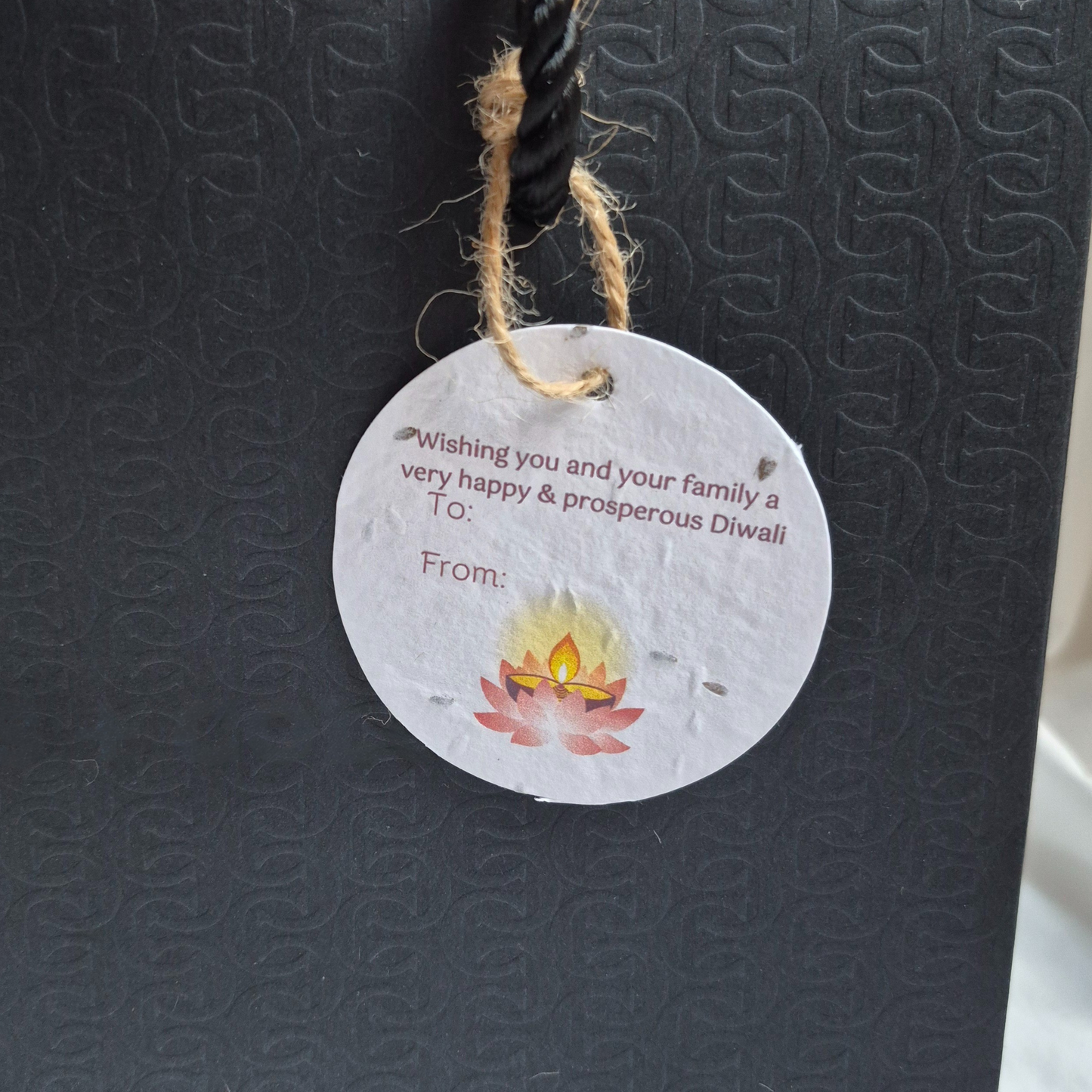 Round Diwali gift tag, made of seed paper, with 'wishing you and your family a happy & prosperous Diwali' text and lotus illustration hanging from a black bag