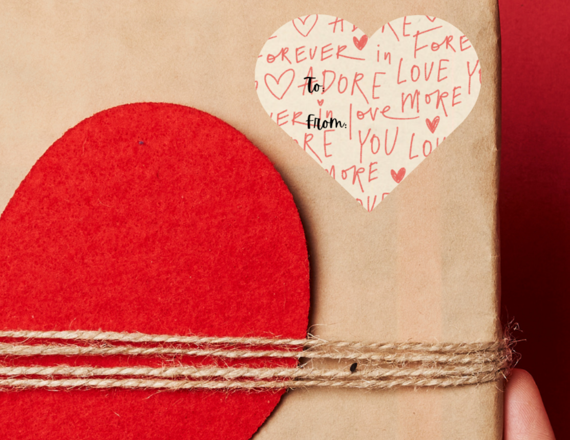 Heart shaped Gift sticker label for Valentine's day, with a lightly printed background that has hearts and 'love you, Adore, and love you forever' placed on a Gift box with red heart design held against a red background
