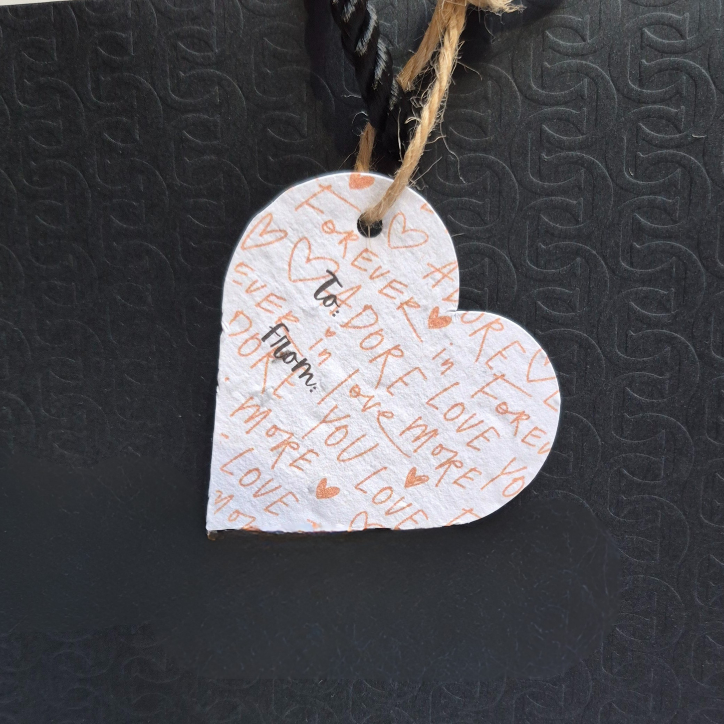 Valentines day heart-shaped seed paper gift tag with a lightly printed background that has hearts and 'love you, Adore, and love you forever' text. Hanging from a black bag.