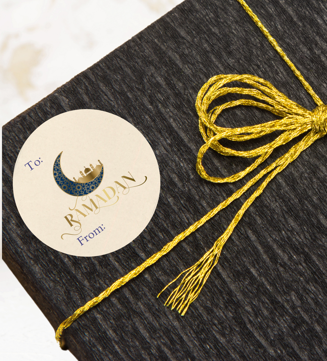 Dark textured box with a gold ribbon and a 'Happy Ramadan 'Moon and Mosque gift sticker on a light background