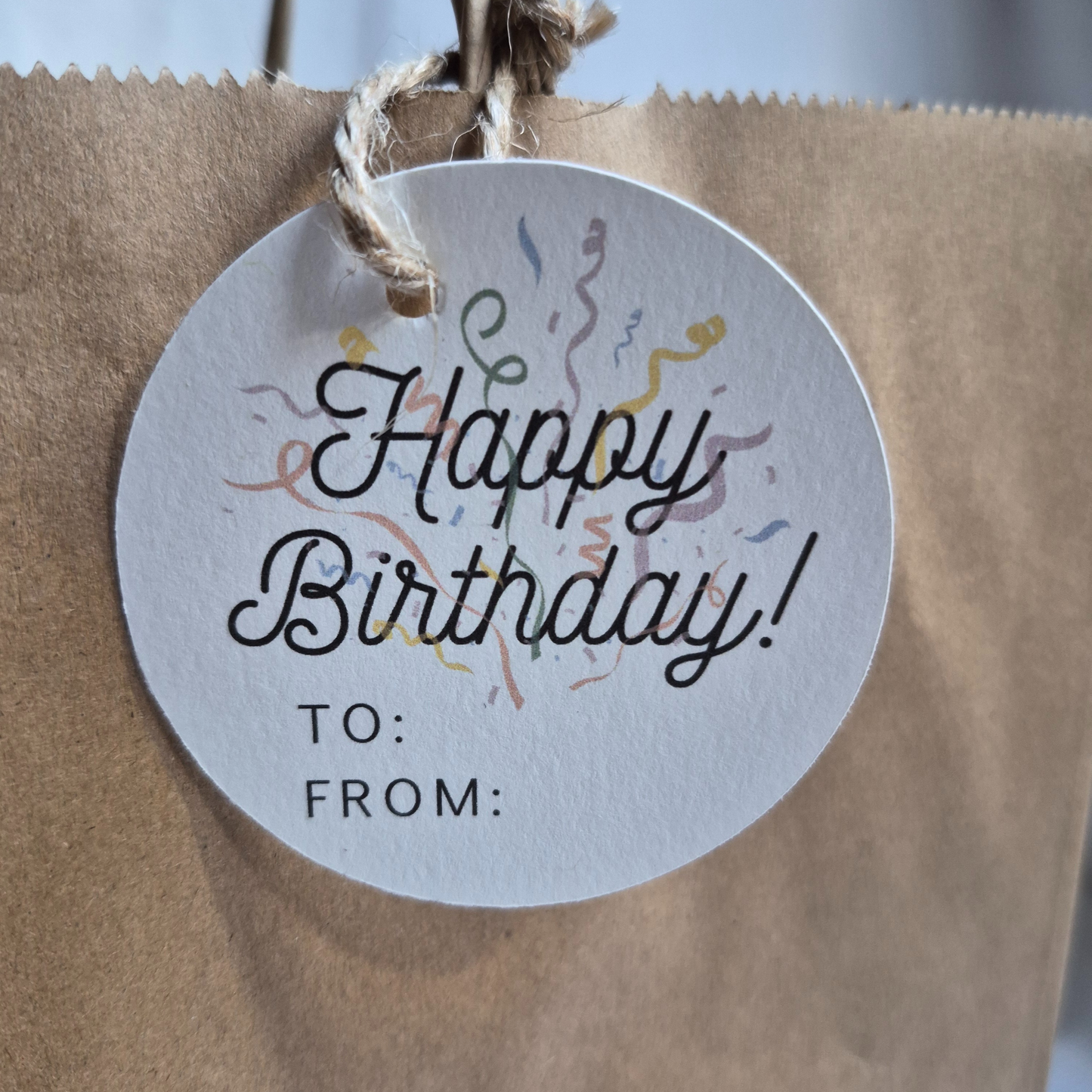 Round Birthday recycled paper gift tag with 'Happy Birthday!' text and confetti on a brown paper bag. Eco-friendly design.