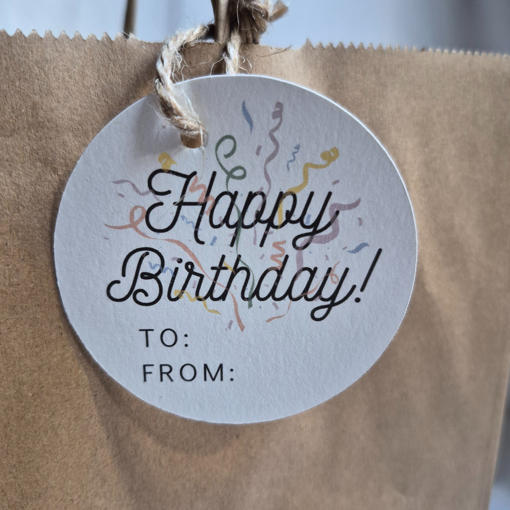 Round Birthday recycled paper gift tag with 'Happy Birthday!' text and confetti on a brown paper bag. Eco-friendly design.