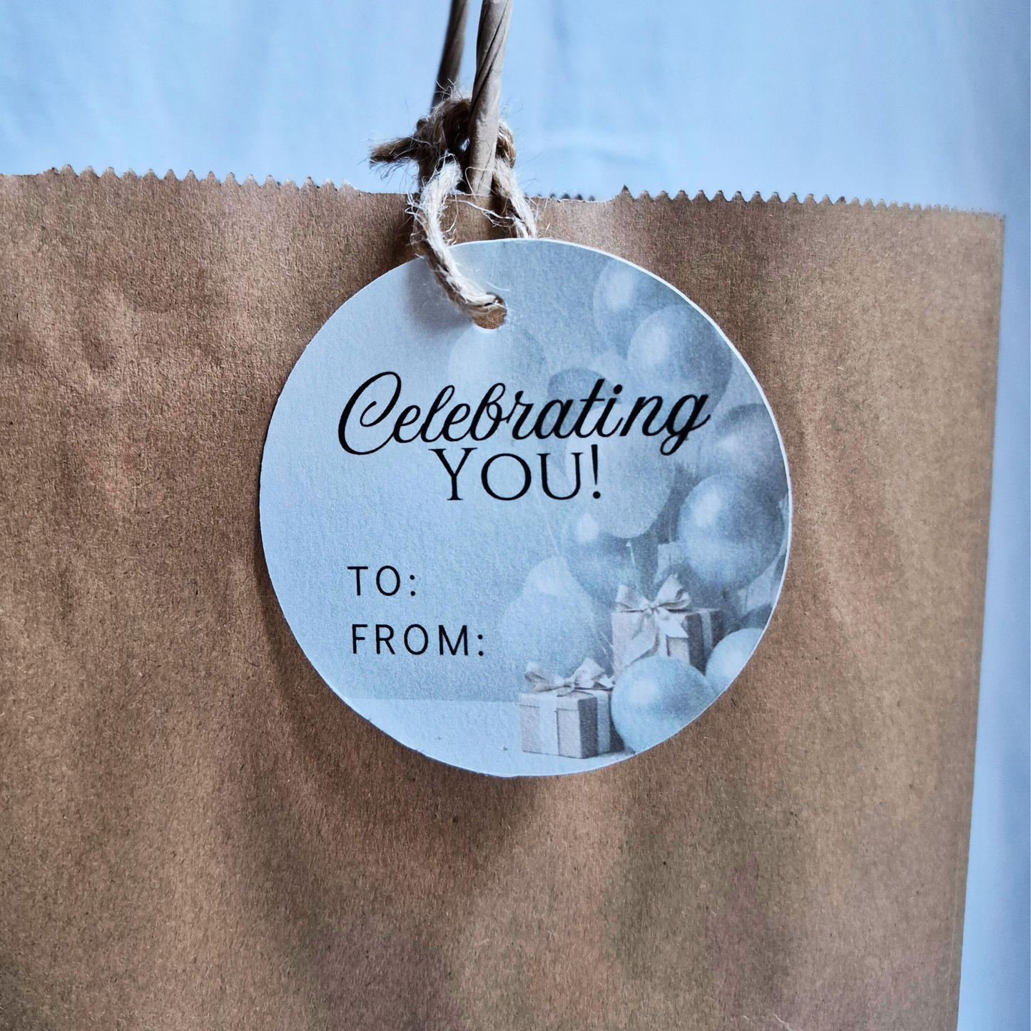 Round  Eco-friendly celebratory gift tag , made with recycled paper, with 'Celebrating YOU!' text and blue balloons on a brown bag