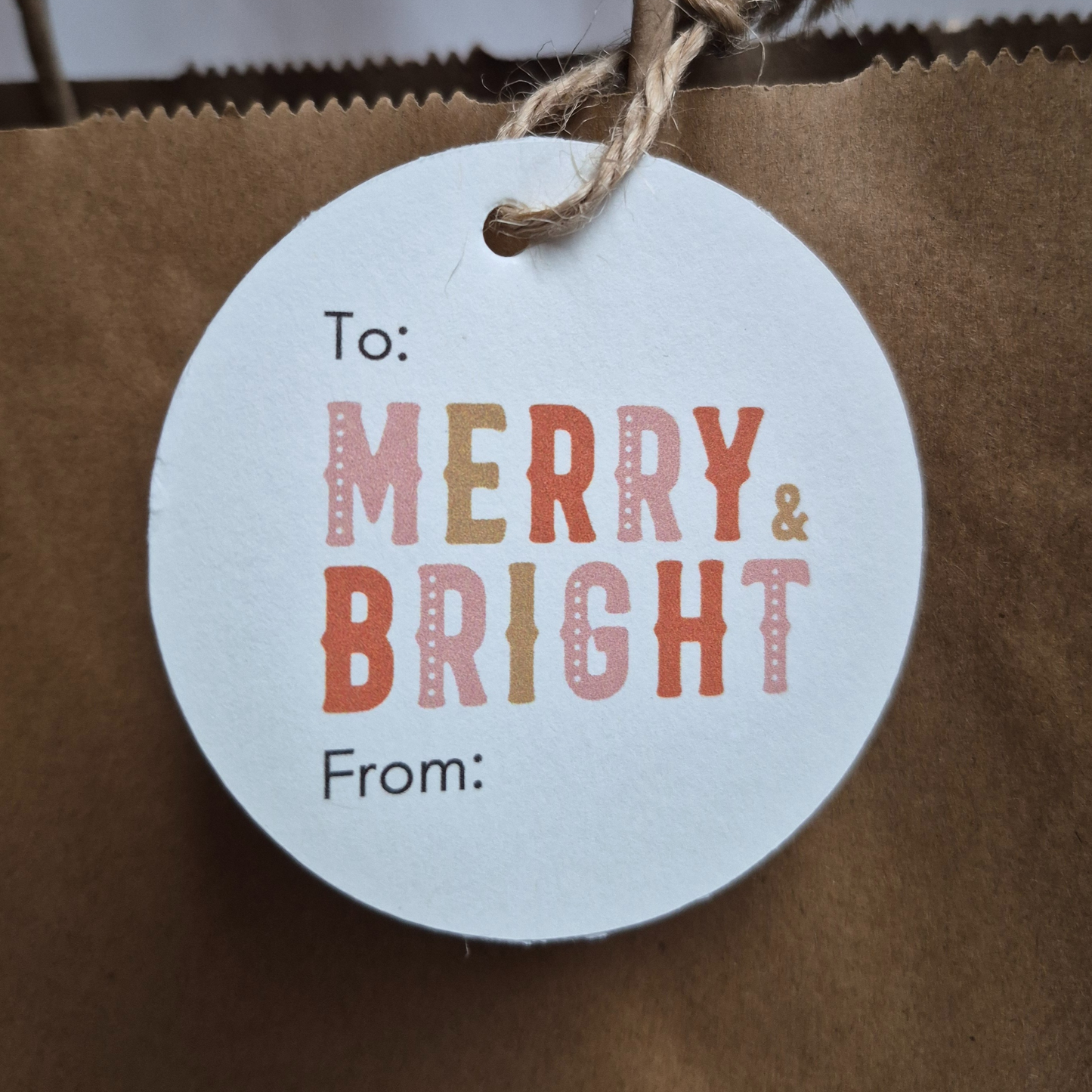 Round Recycled paper Christmas gift tag with 'Merry & Bright' text hanging from a brown paper bag.
