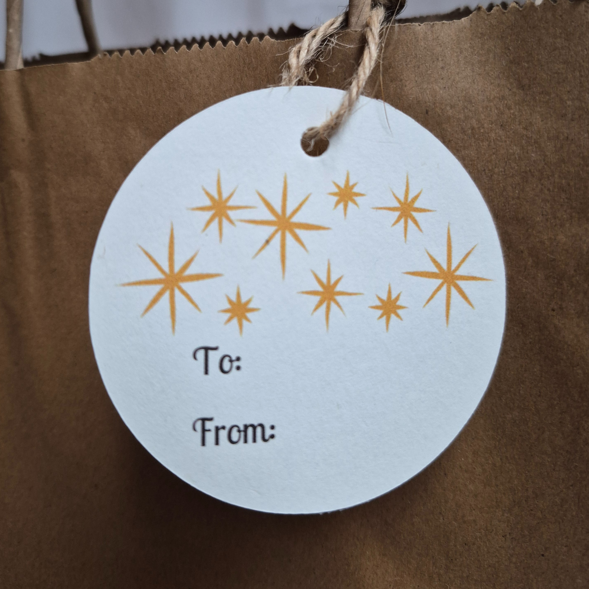 Round Christmas gift tag made of  recycled paper with gold stars hanging from a brown paper bag