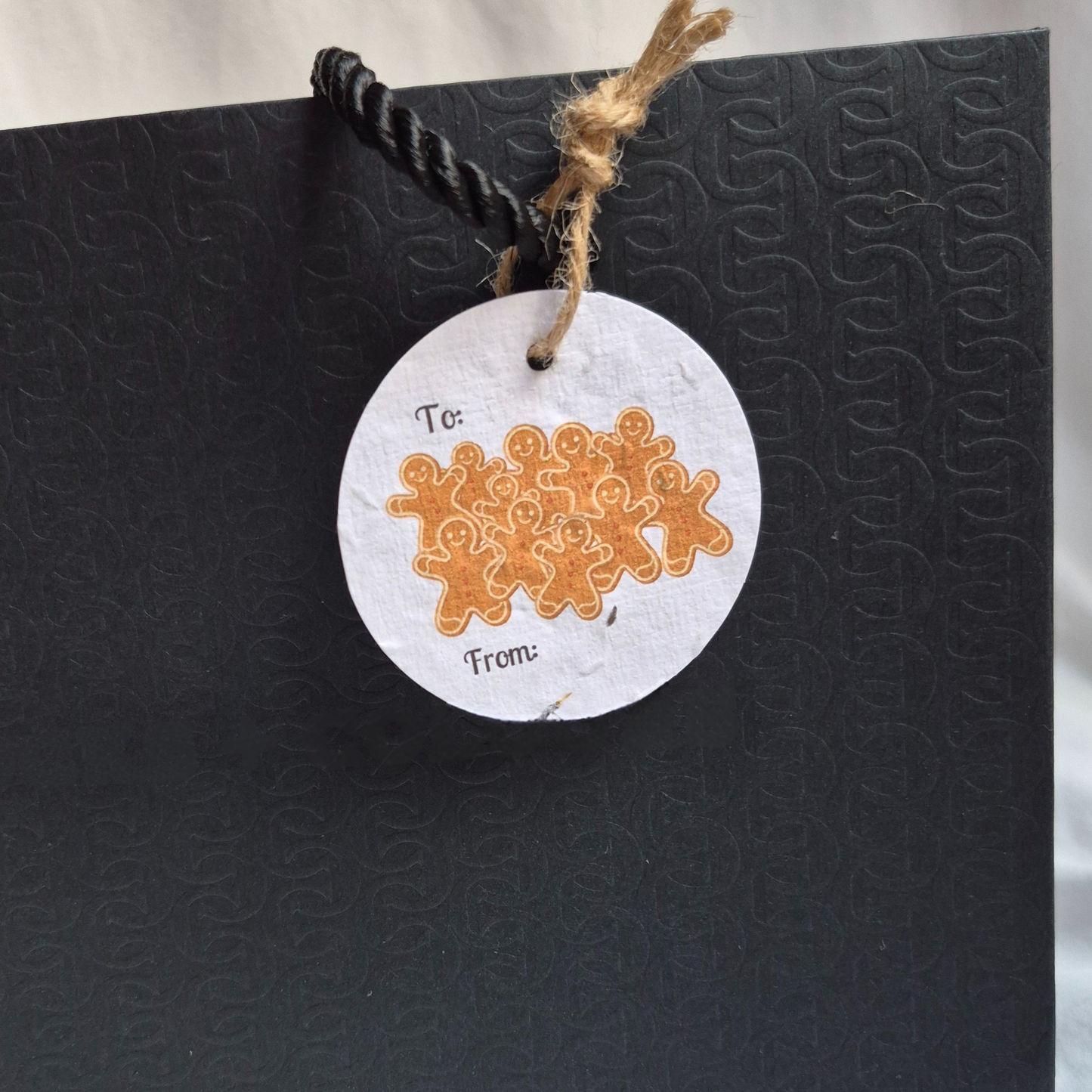 Round Christmas gift tag made from seed paper, with 'a pile of gingerbread men illustration, hanging from a black bag.