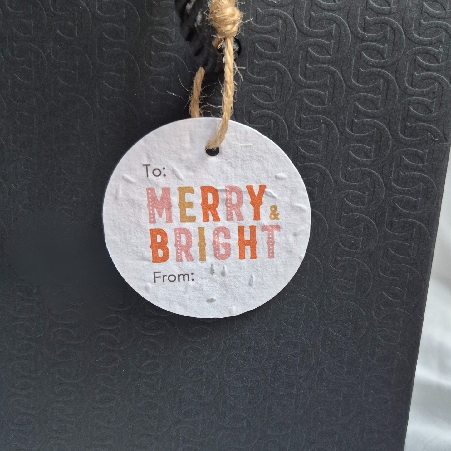 Round Christmas gift tag made from seed paper, with 'Merry & Bright' text hanging from a black bag.