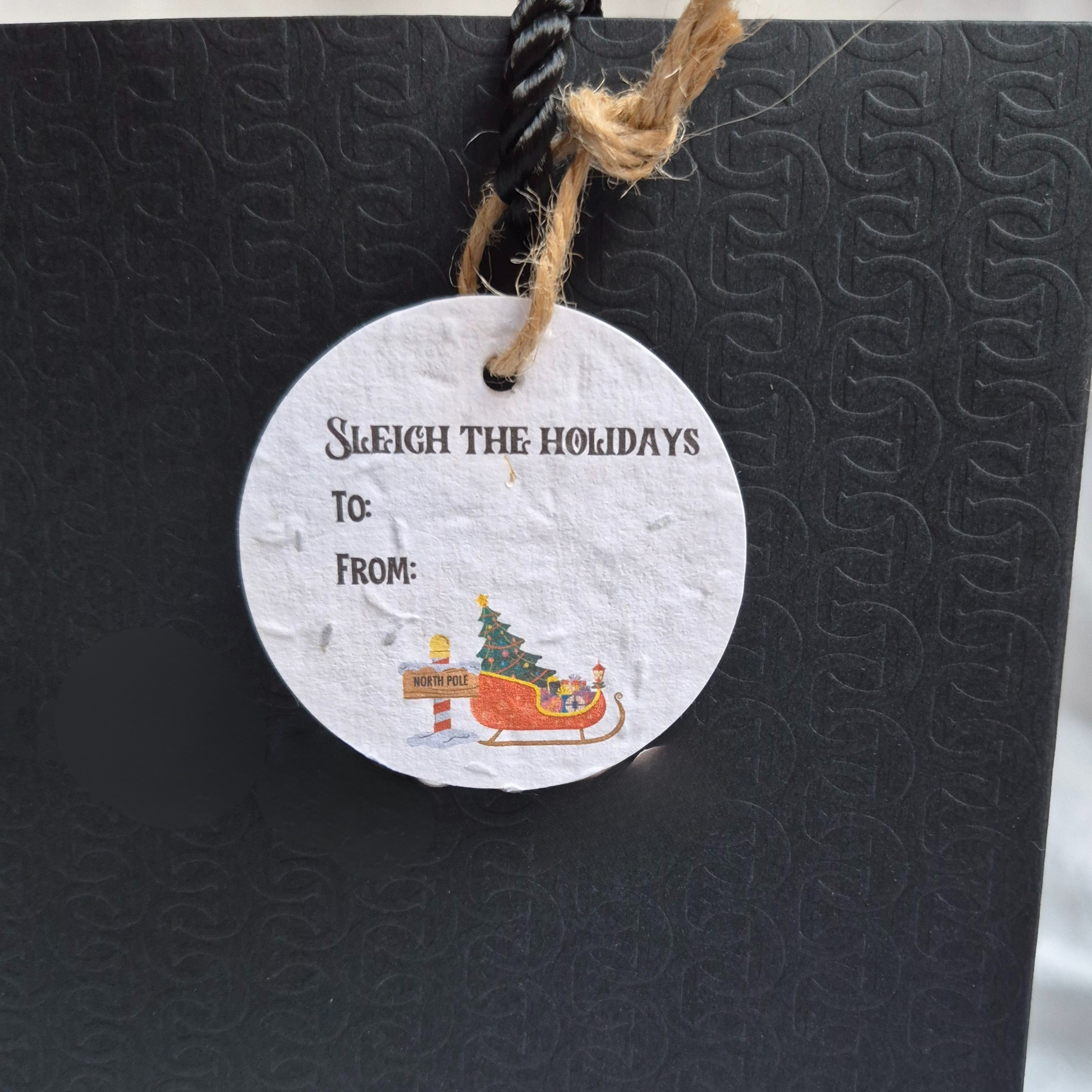 Round Christmas gift tag made from seed paper, with 'Sleigh the Holidays' text and a Santa sleigh in the North Pole illustration,hanging from a black bag.