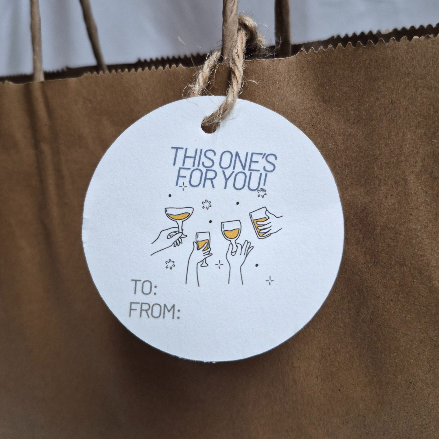 Round recycled paper gift tag with 'This one's for you!' text and wine glass illustrations on a brown paper bag.