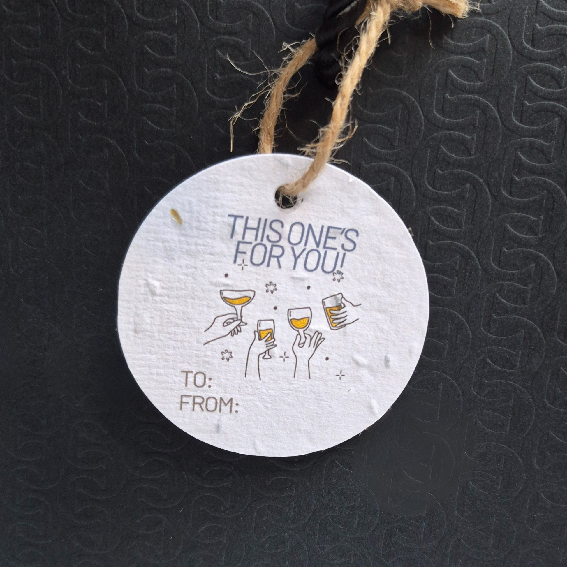 Round seed paper gift tag with 'This one's for you!' text and wine glass illustrations on a black textured background.