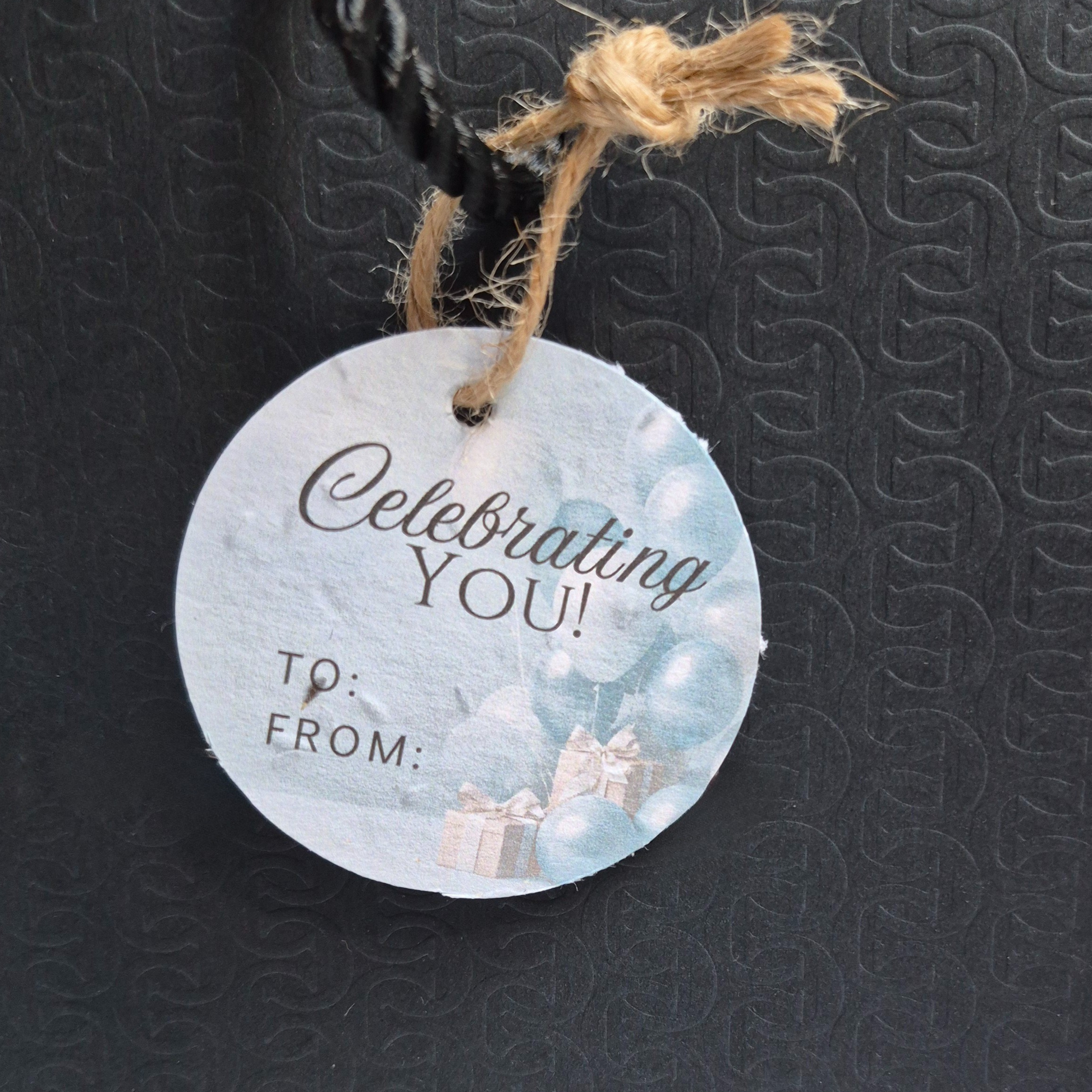 Round seed paper gift tag with 'Celebrating You!' text and balloons on a black gift bag