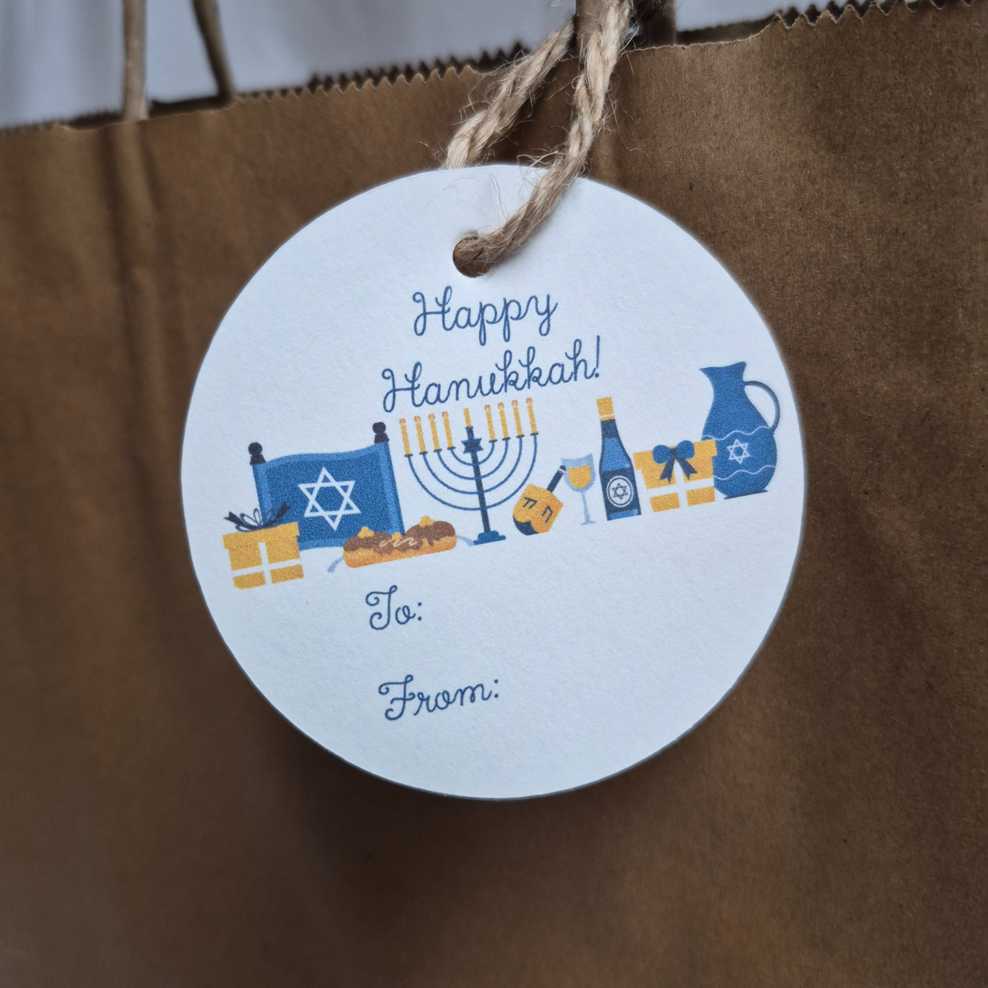 Round Hanukkah gift tag with 'Happy Hanukkah' text and table setting illustration on a brown paper bag.