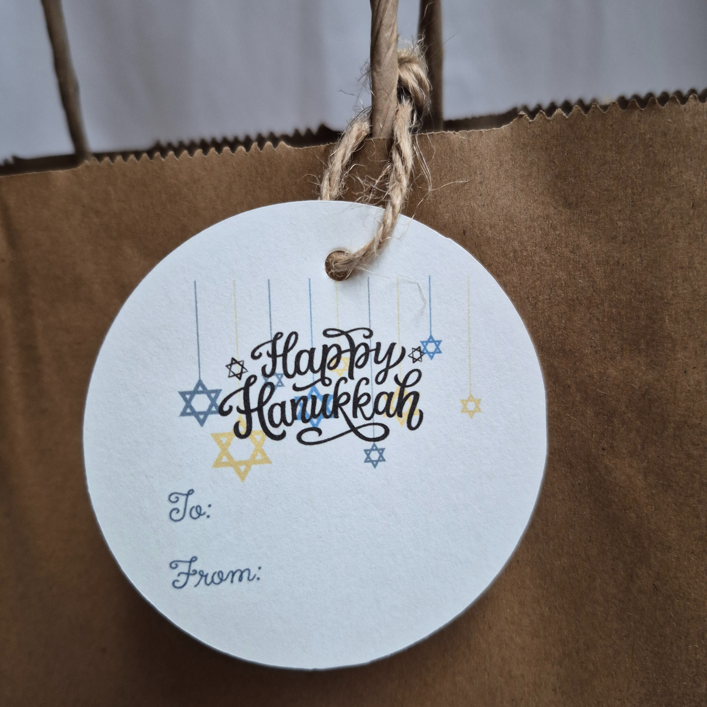 Round Hanukkah gift tag with 'Happy Hanukkah' text and star of David designs hanging from a brown paper bag.