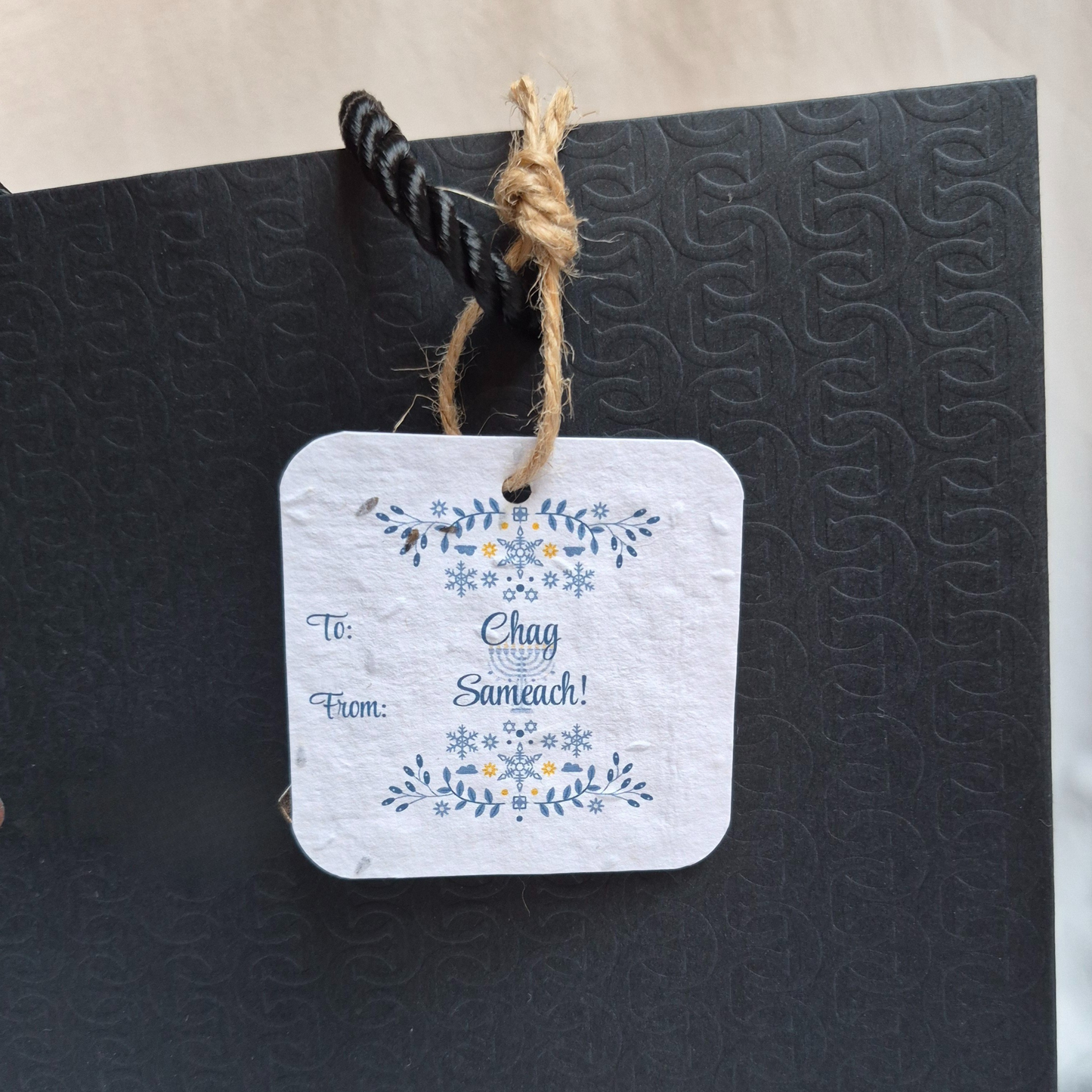 Square Hanukkah seed paper gift tag with Chag Sameach!' text. Hanging from a black bag.