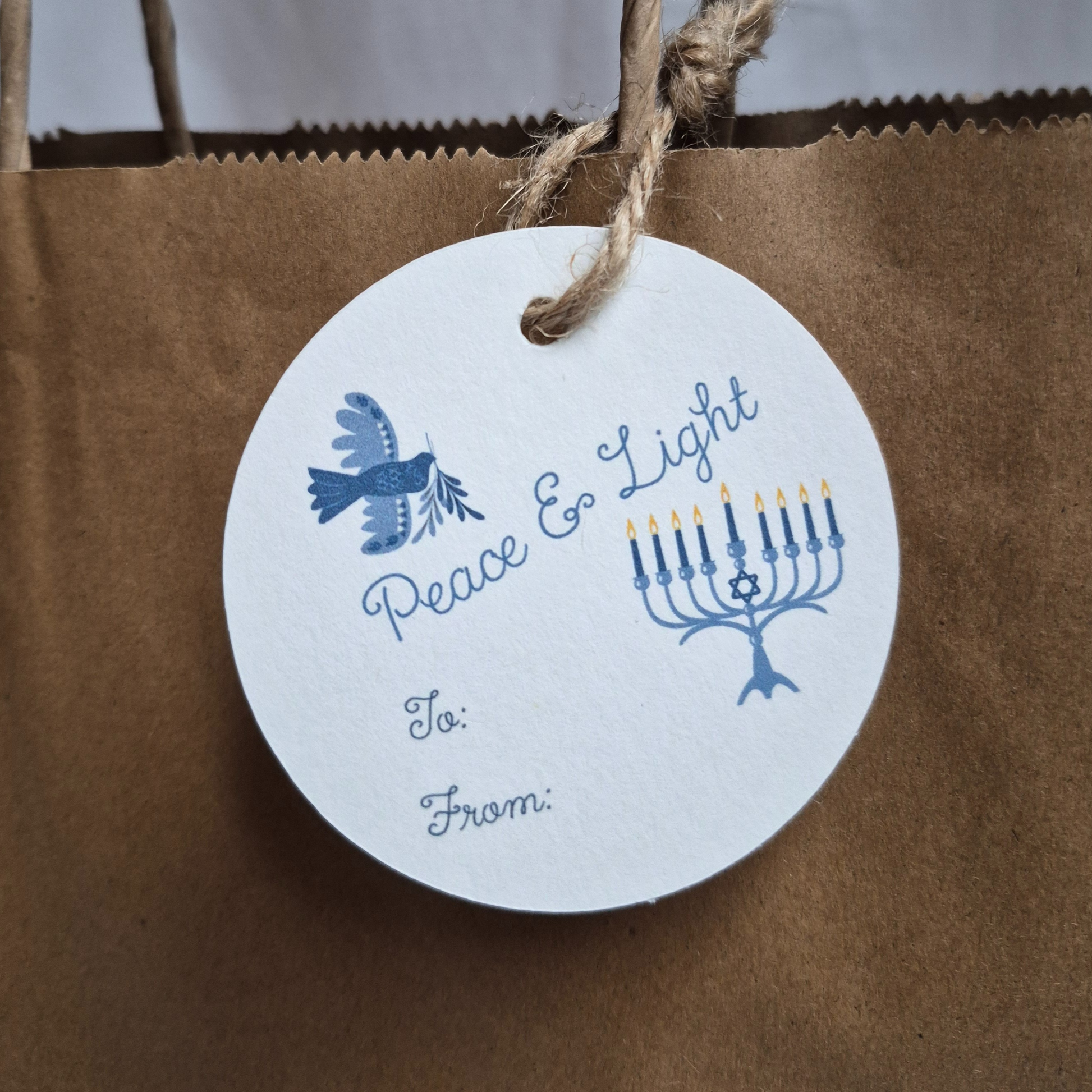 Round Hanukkah gift tag with 'Peace & Light' text and Menorah design on a brown paper bag.