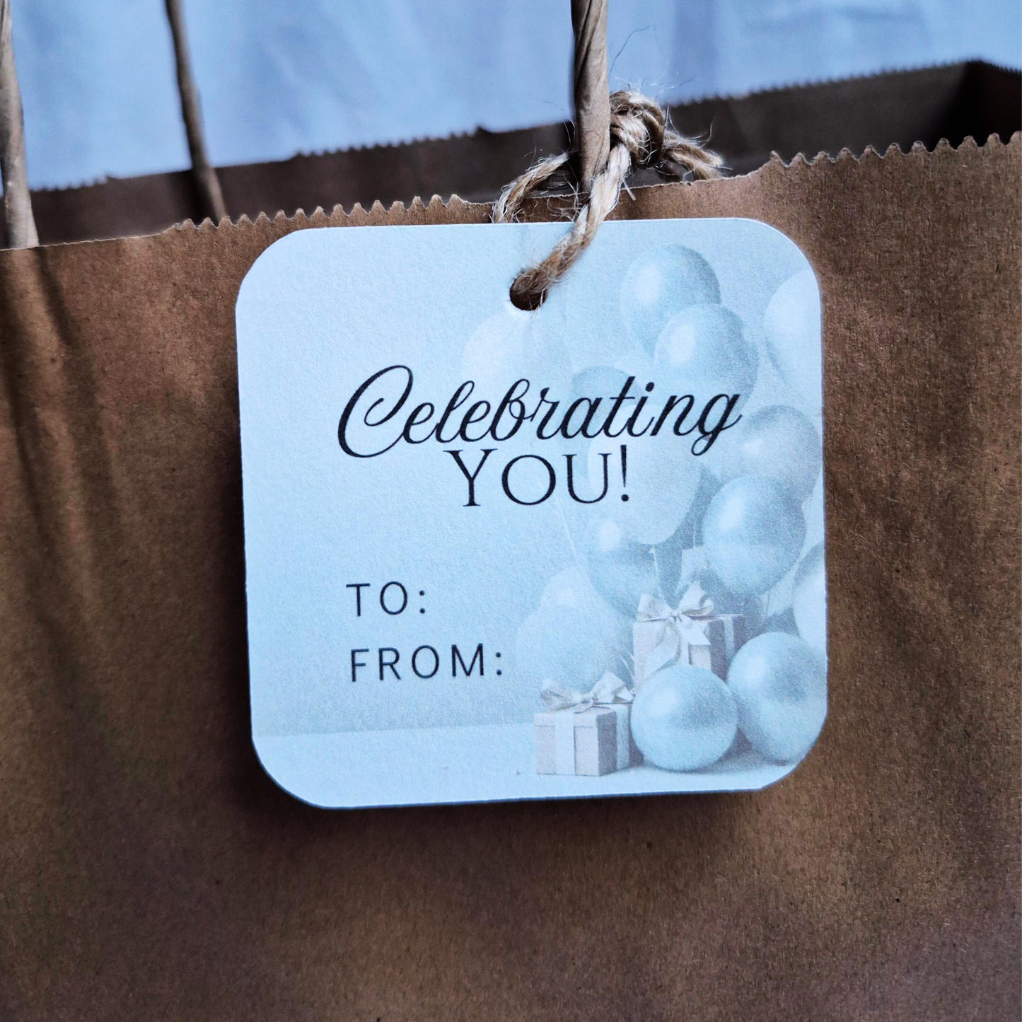 Square Birthday gift tag with 'Celebrating YOU!' text and blue balloons, made of recycled paper, eco-friendly.  Hanging from a brown bag