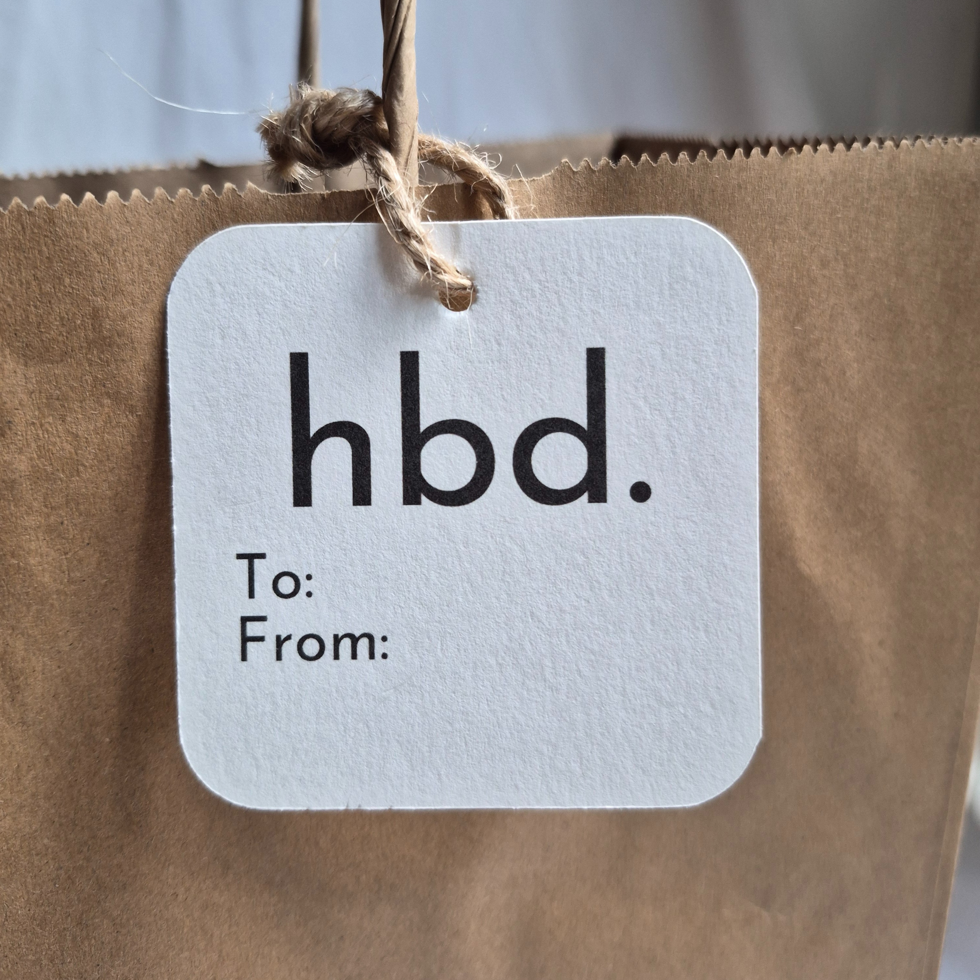 Square Birthday gift tag with 'hbd.'  text, made of recycled paper. Hanging from a brown bag