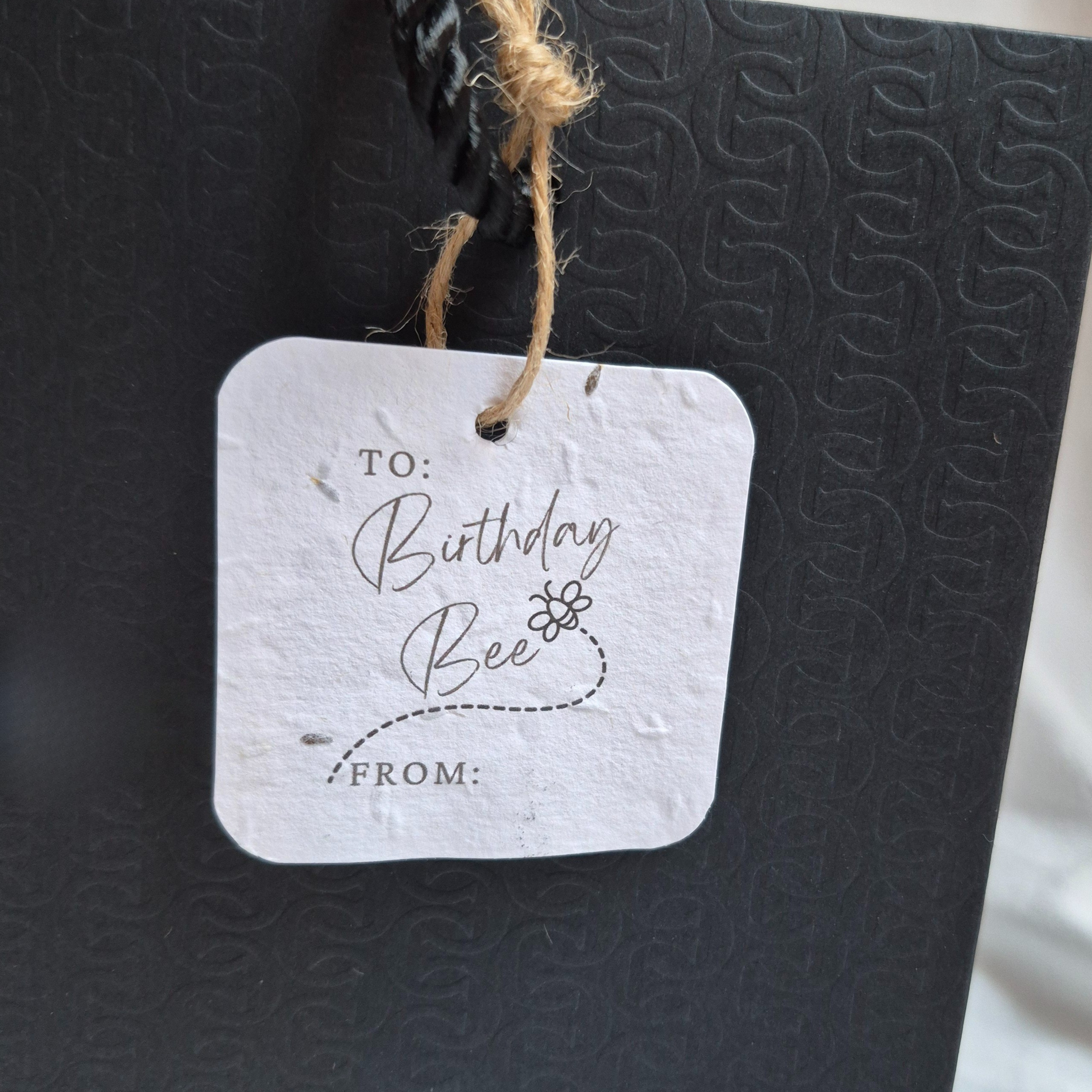 Square Birthday plantable gift tag with 'Birthday Bee' text  and a bee illustration , made of seed paper. Hanging from a black bag