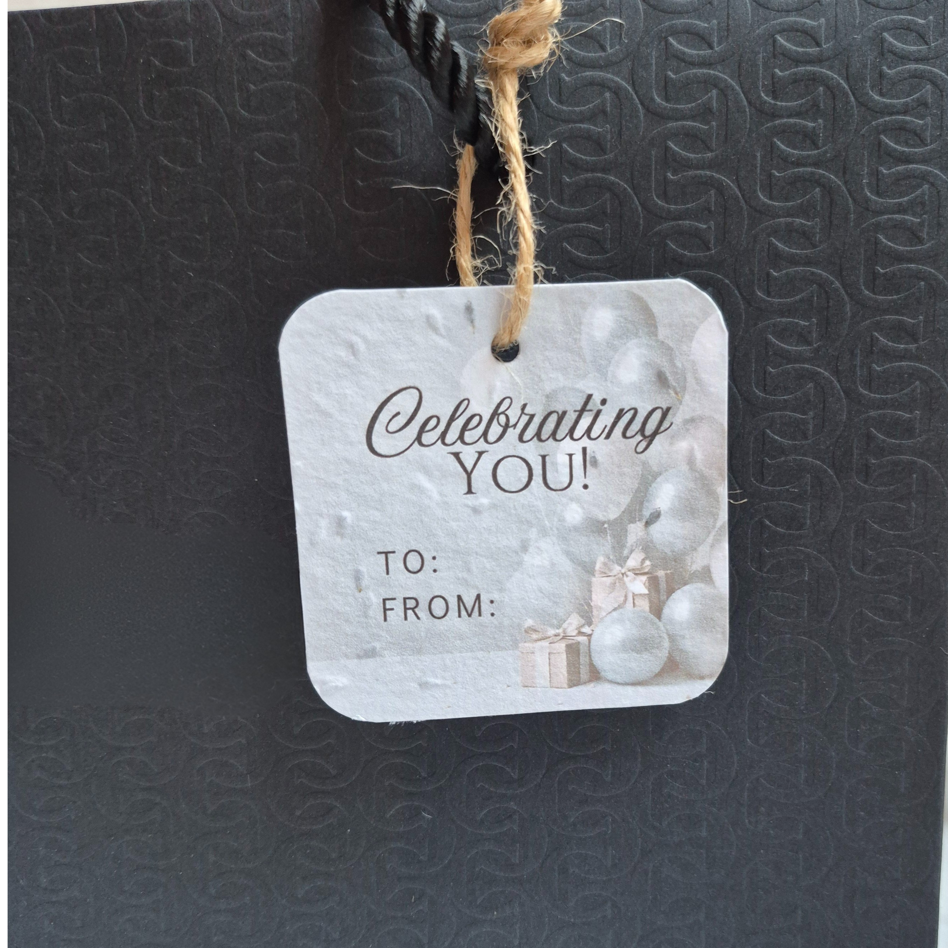 Square Birthday plantable gift tag with 'Celebrating YOU!' text and blue balloons, made of seed paper. Hanging from a black bag