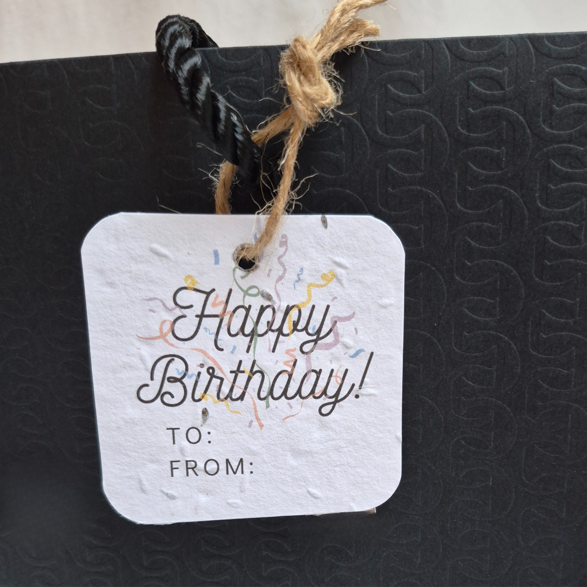 Square Birthday plantable gift tag with 'Happy Birthday!' text and confetti , made of seed paper. Hanging from a black bag