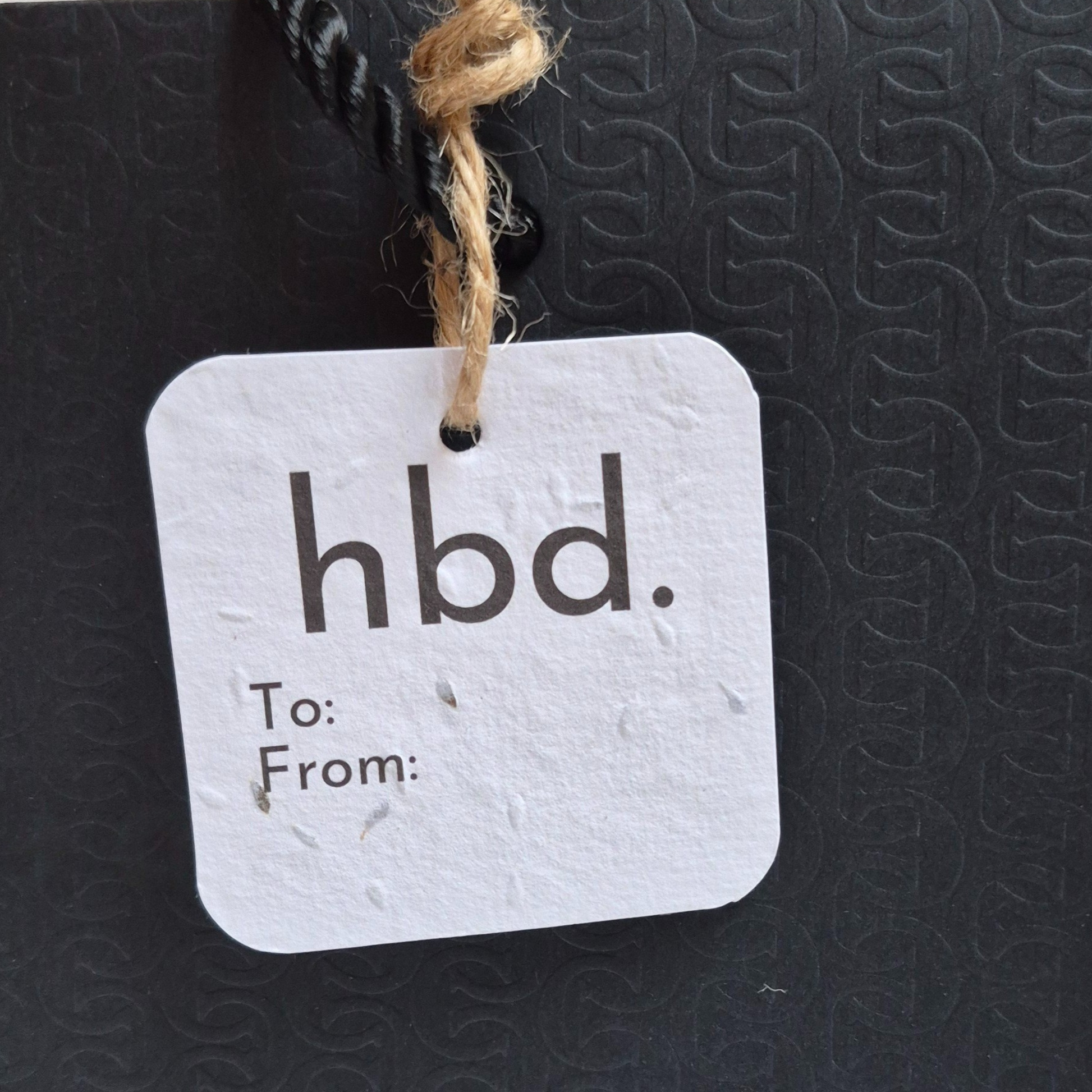Square Birthday plantable gift tag with 'hbd.' text, made of seed paper. Hanging from a black bag