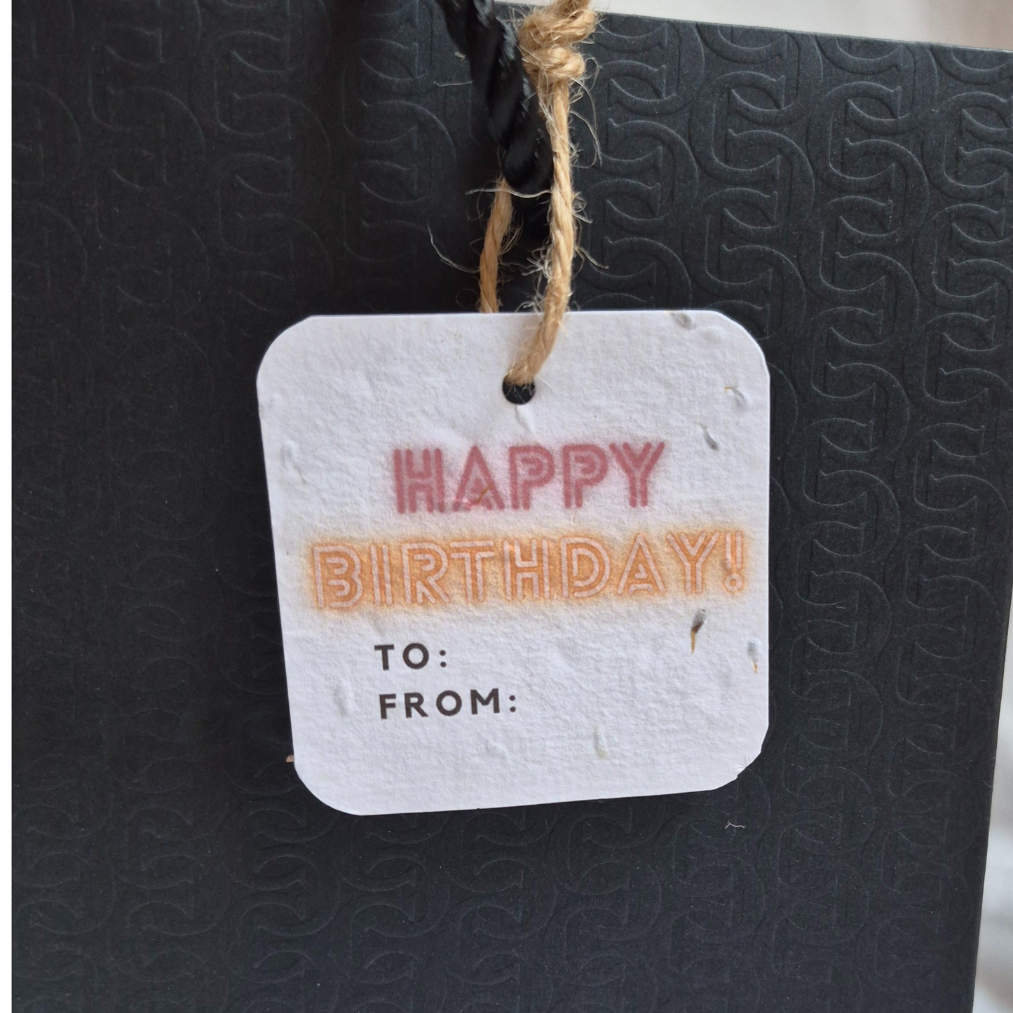 Square Birthday plantable gift tag with 'Happy Birthday!' in neon text, made of seed paper. Hanging from a black bag