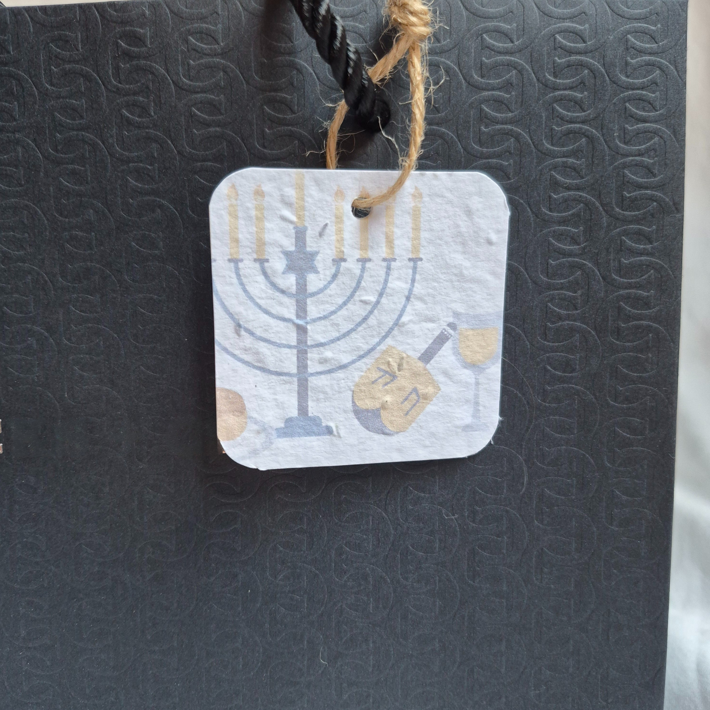 Square Hanukkah seed paper gift tag with Menorah and dreidel design. Hanging from a black bag.