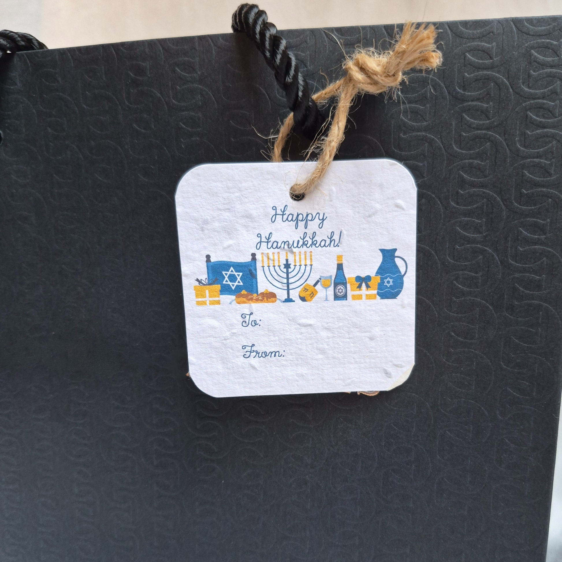 Square Hanukkah seed paper gift tag with 'Happy Hanukkah' text and table setting illustration. Hanging from a black bag.