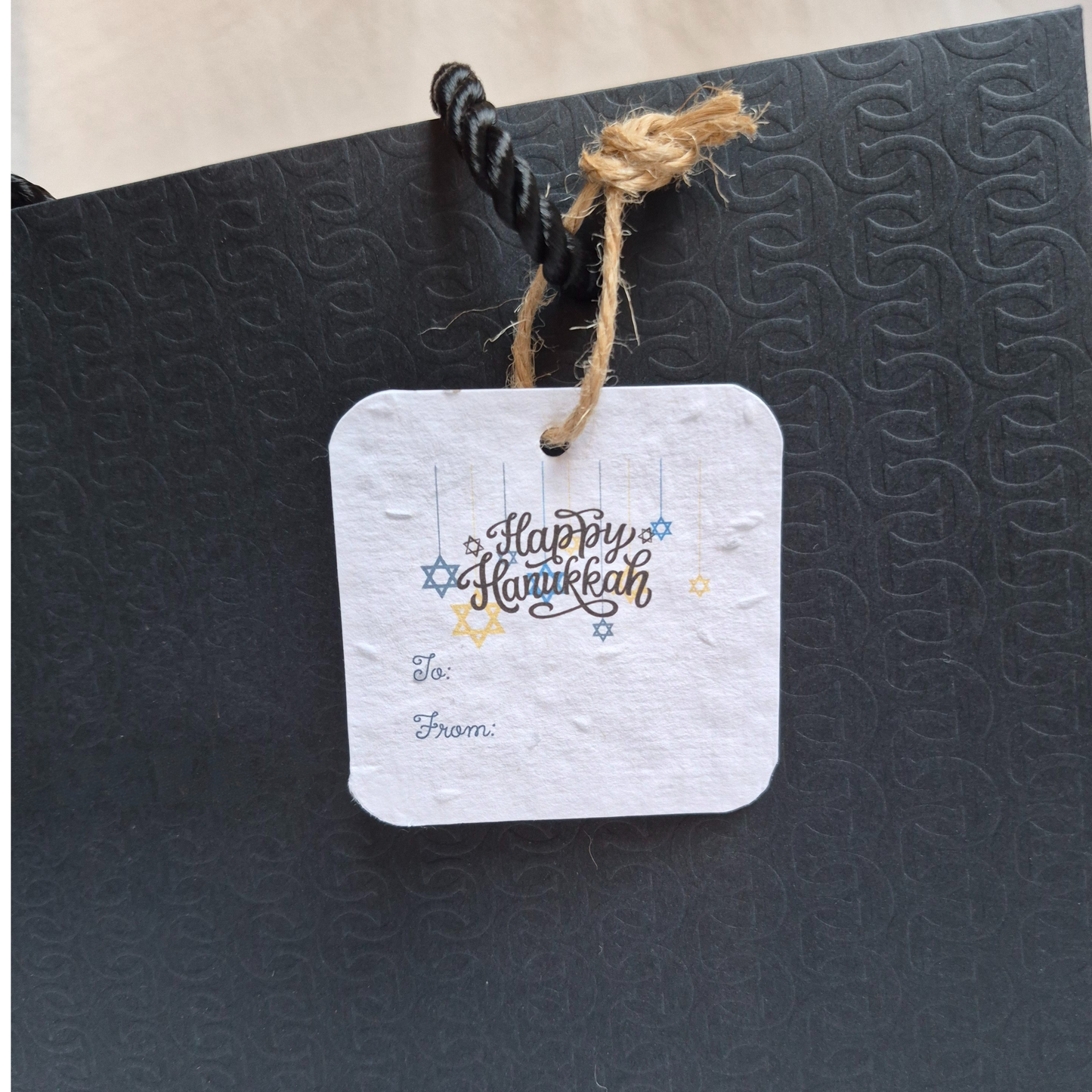 Square Hanukkah seed paper gift tag with 'Happy Hanukkah' text and star of David designs. Hanging from a black bag.