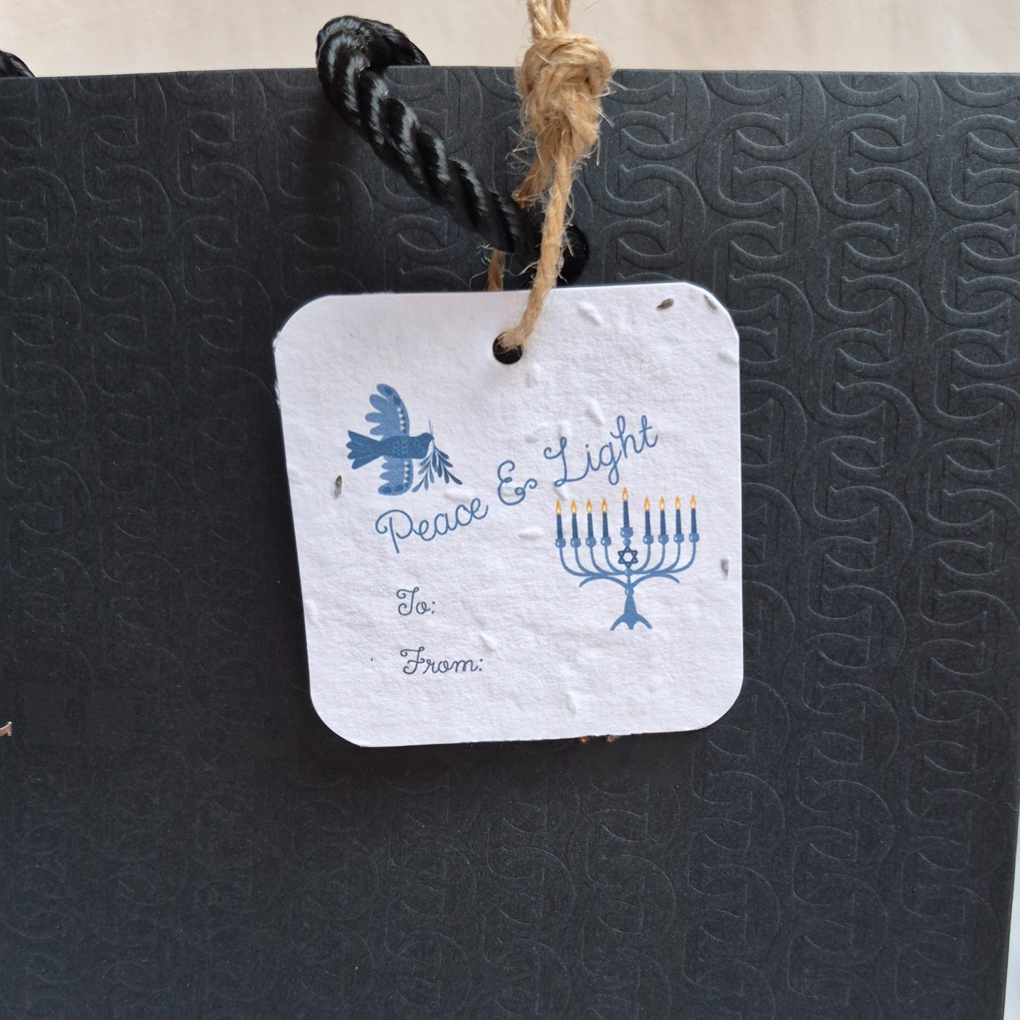 Square Hanukkah seed paper gift tag with 'Peace & Light' text and Menorah design. Hanging from a black bag.