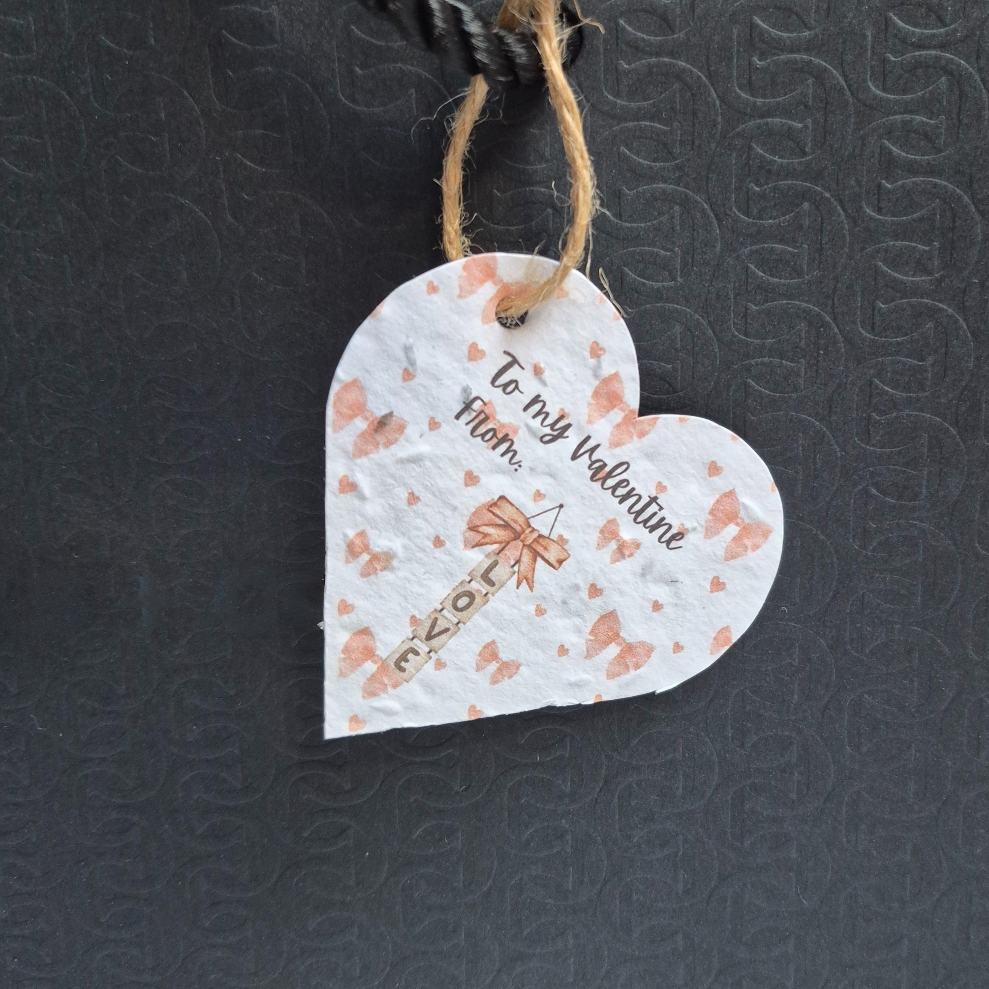 Valentine's day  heart-shaped plantable seed paper gift tag with pink bows in the background and 'To my Valentine' text. and 'LOVE' hanging vertically. Hanging from a  black bag