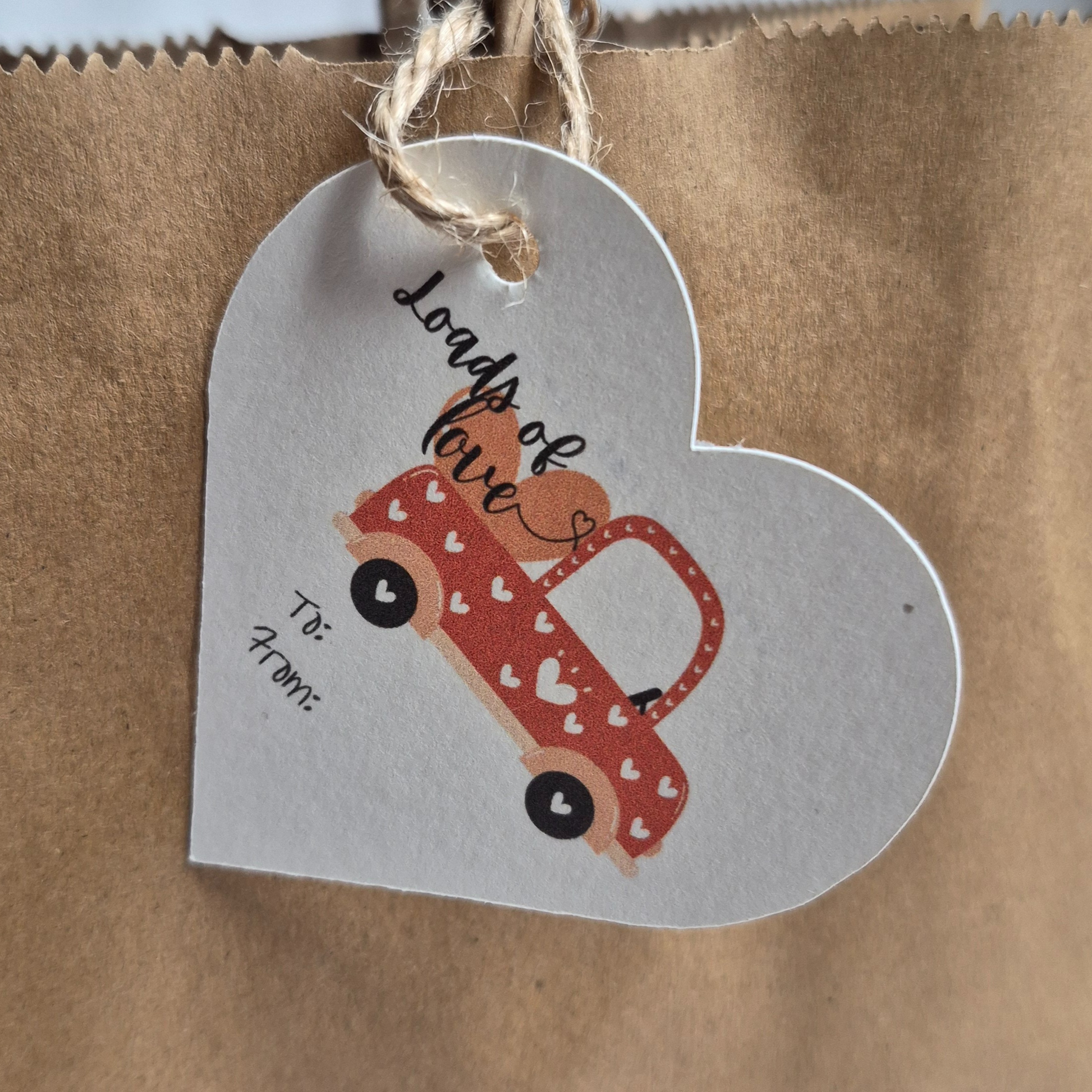 Heart-shaped  Valentines day gift tag with a red truck with a truckload filled with 'loads of love' text. Hanging from a brown paper bag.